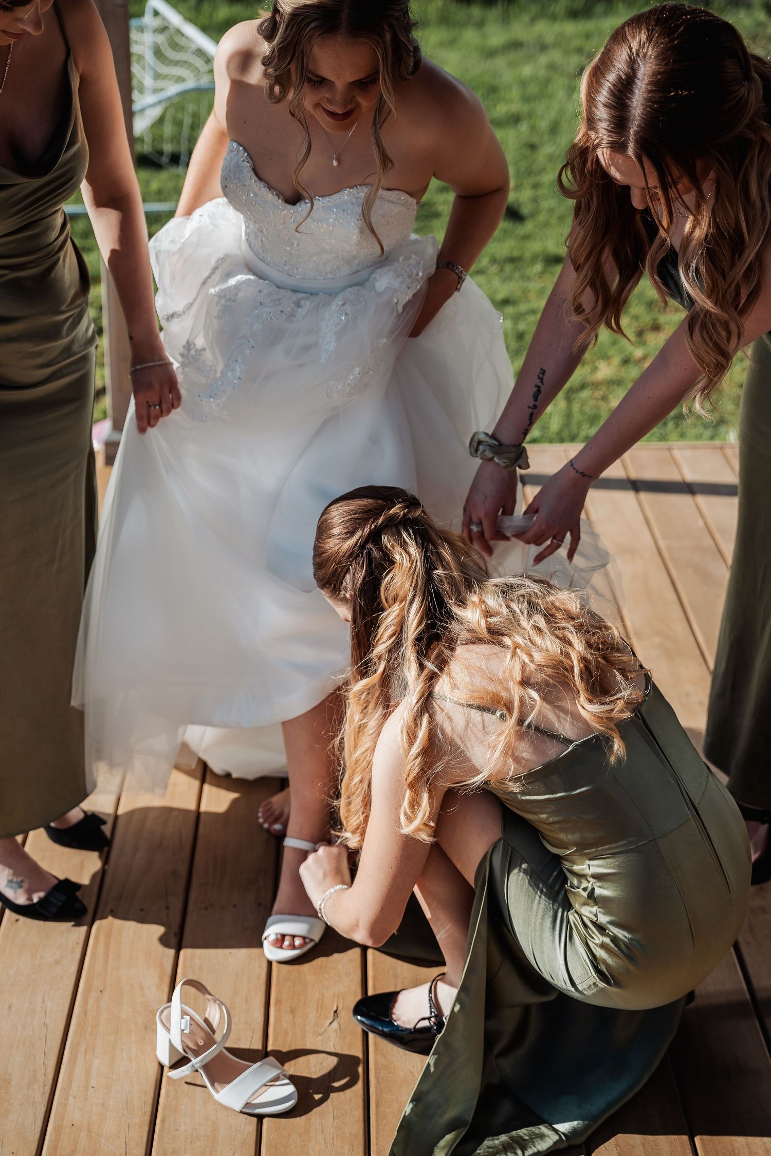 Bridesmaids help a bride adjust her shoes on wooden deck during outdoor wedding preparations.