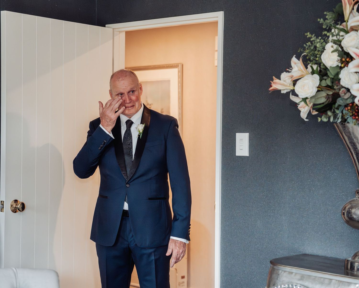 Emotional man in navy suit wiping tears at wedding ceremony, standing in doorway with floral arrangement nearby.