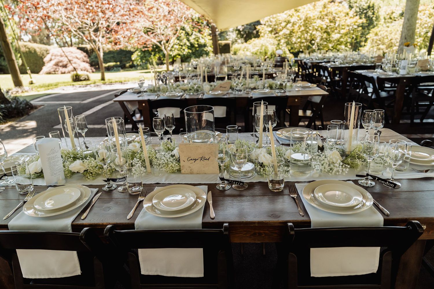 Elegant outdoor wedding reception table set with white floral centerpieces, fine china, and glassware under a canopy.