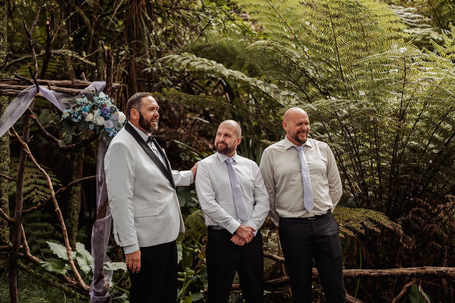 Three groomsmen stand together in a lush forest wedding ceremony backdrop with fern trees.