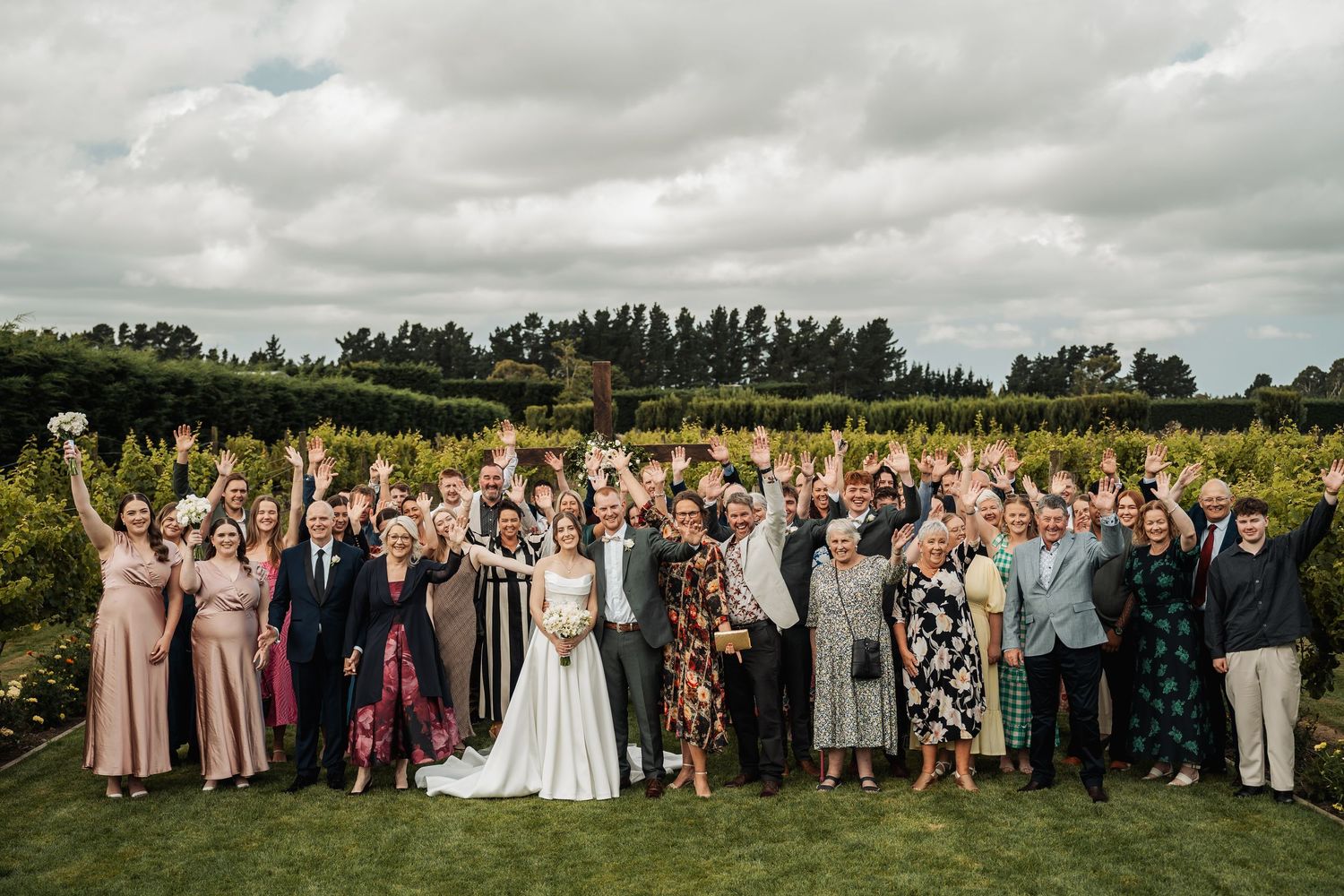 Large wedding party celebrates outdoors in vineyard garden with arms raised joyfully under cloudy sky.