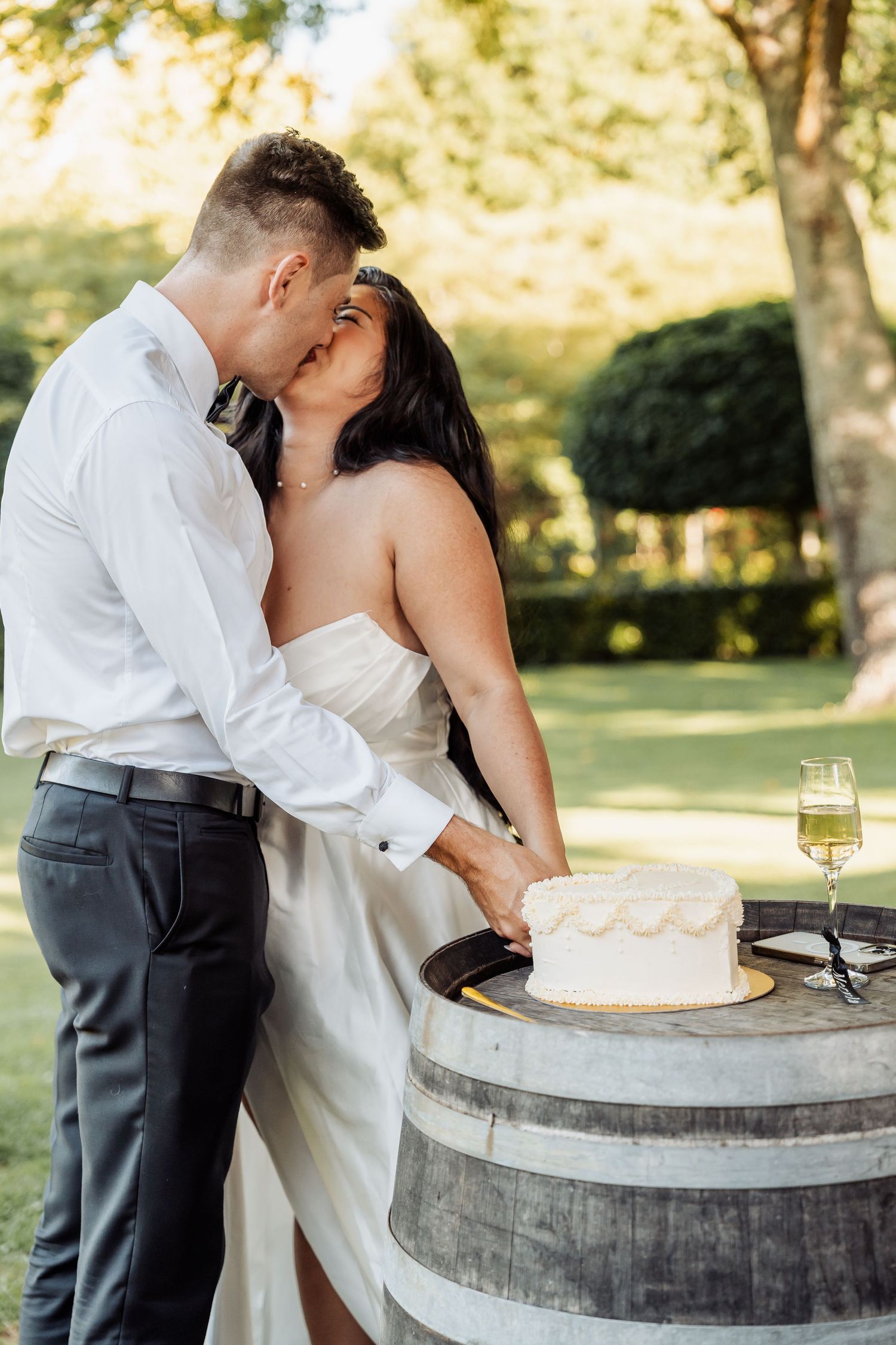 Bride and groom kissing while cutting their white wedding cake on a wooden barrel outdoors.