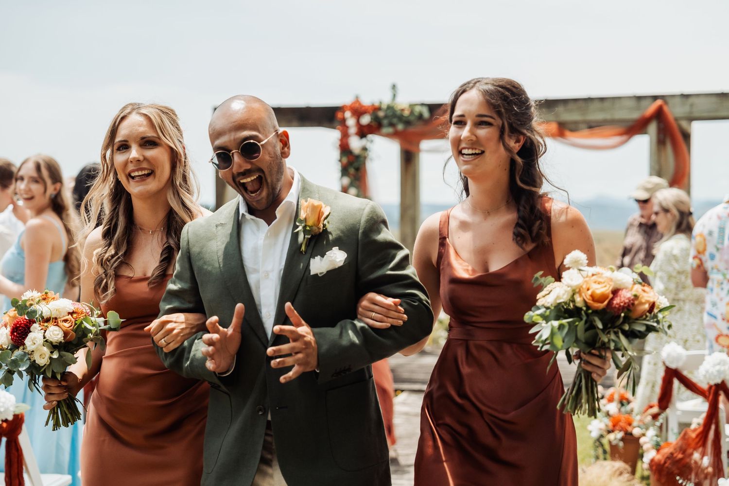 Groom in green suit walks down aisle with two bridesmaids in rust dresses holding bouquets.