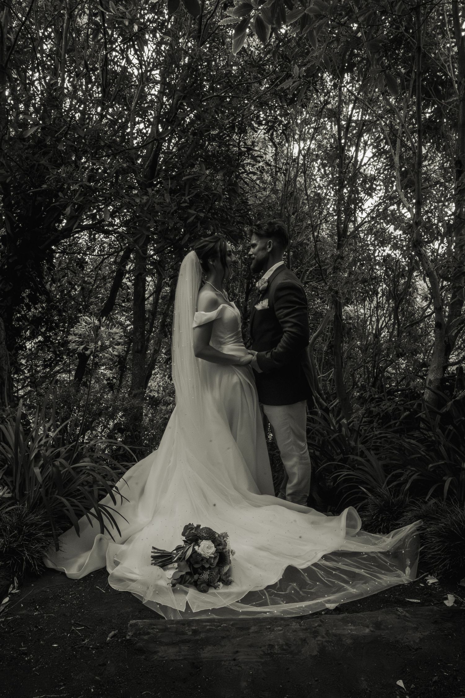 Black and white romantic photo of bride and groom kissing in a lush forest setting.