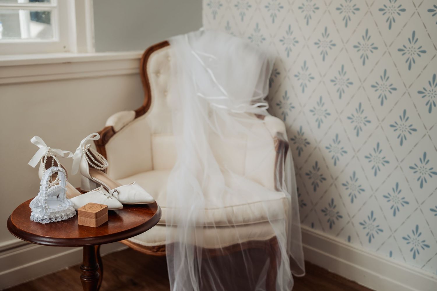 A white wedding dress draped over an elegant vintage chair beside bridal heels on a wooden table, set against floral wallpaper.