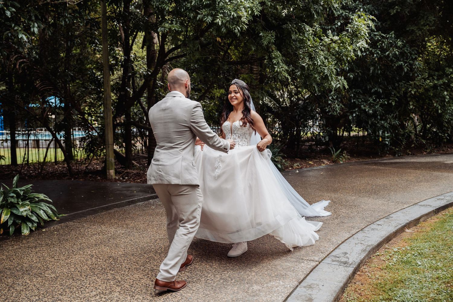 Bride in white gown twirls with groom in gray suit on garden path surrounded by lush green trees.