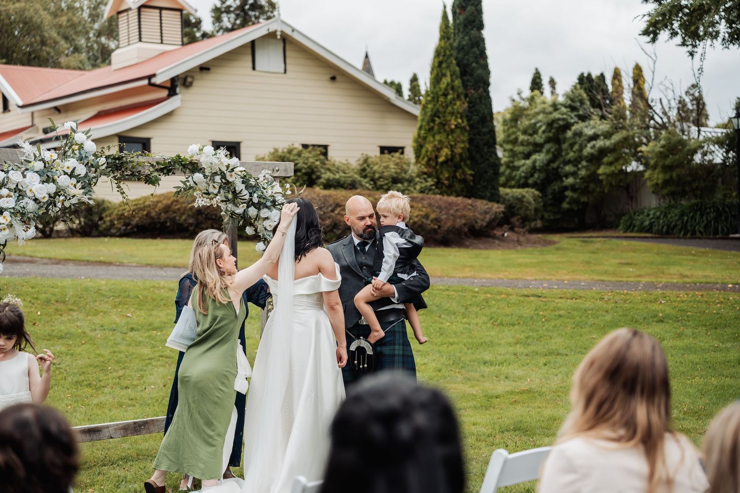 Bride and groom celebrate outdoors at a garden wedding ceremony, with guests seated and a charming white chapel in the background.