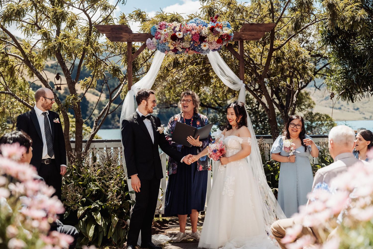 Bride and groom celebrate under a floral arch with officiant during outdoor wedding ceremony.