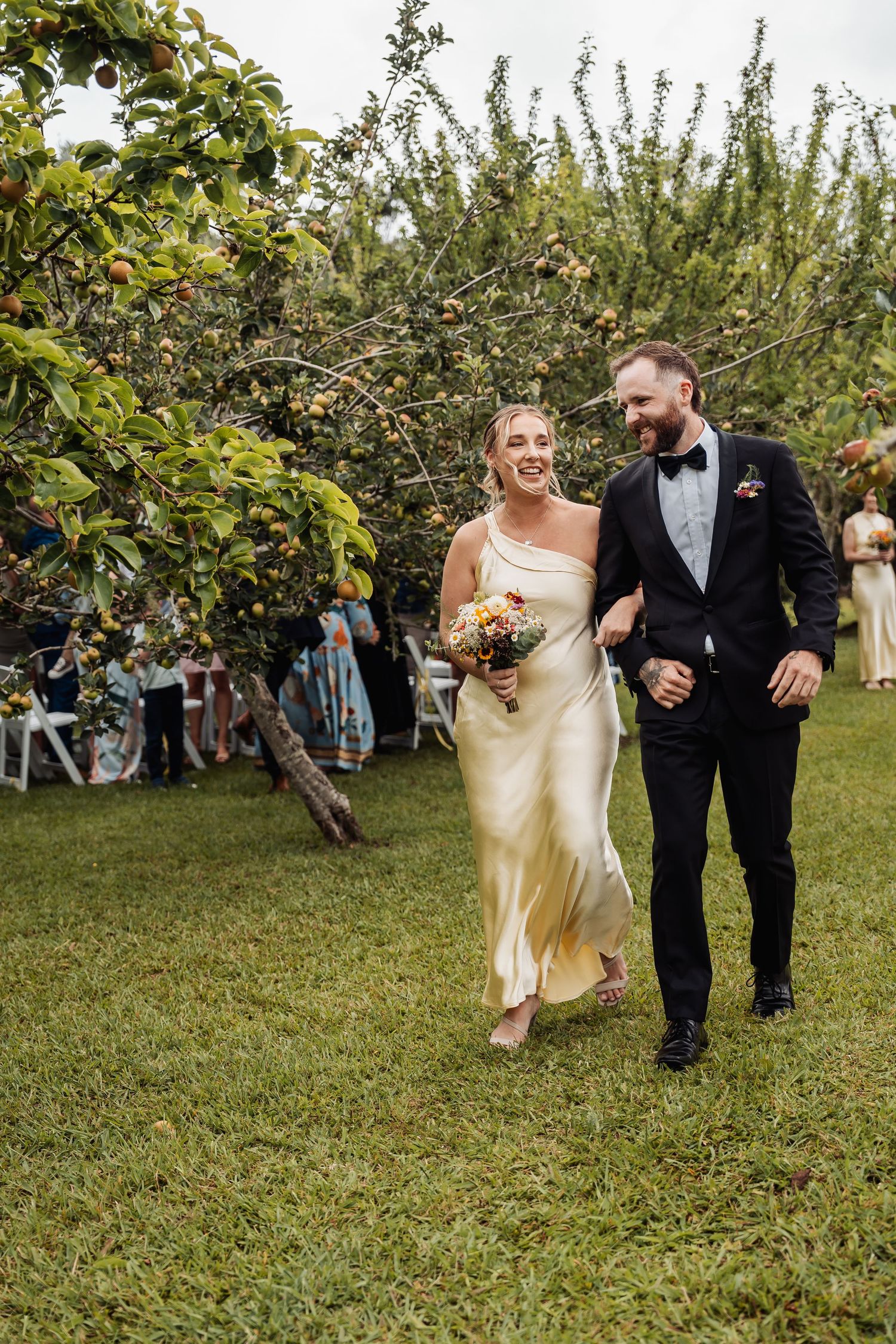 Smiling bride in gold satin gown and groom in black tuxedo walking through a garden.