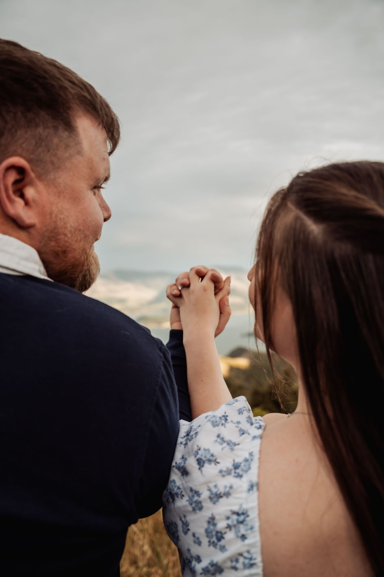 Couple holding hands tenderly against a cloudy sky backdrop during an outdoor engagement photo session.