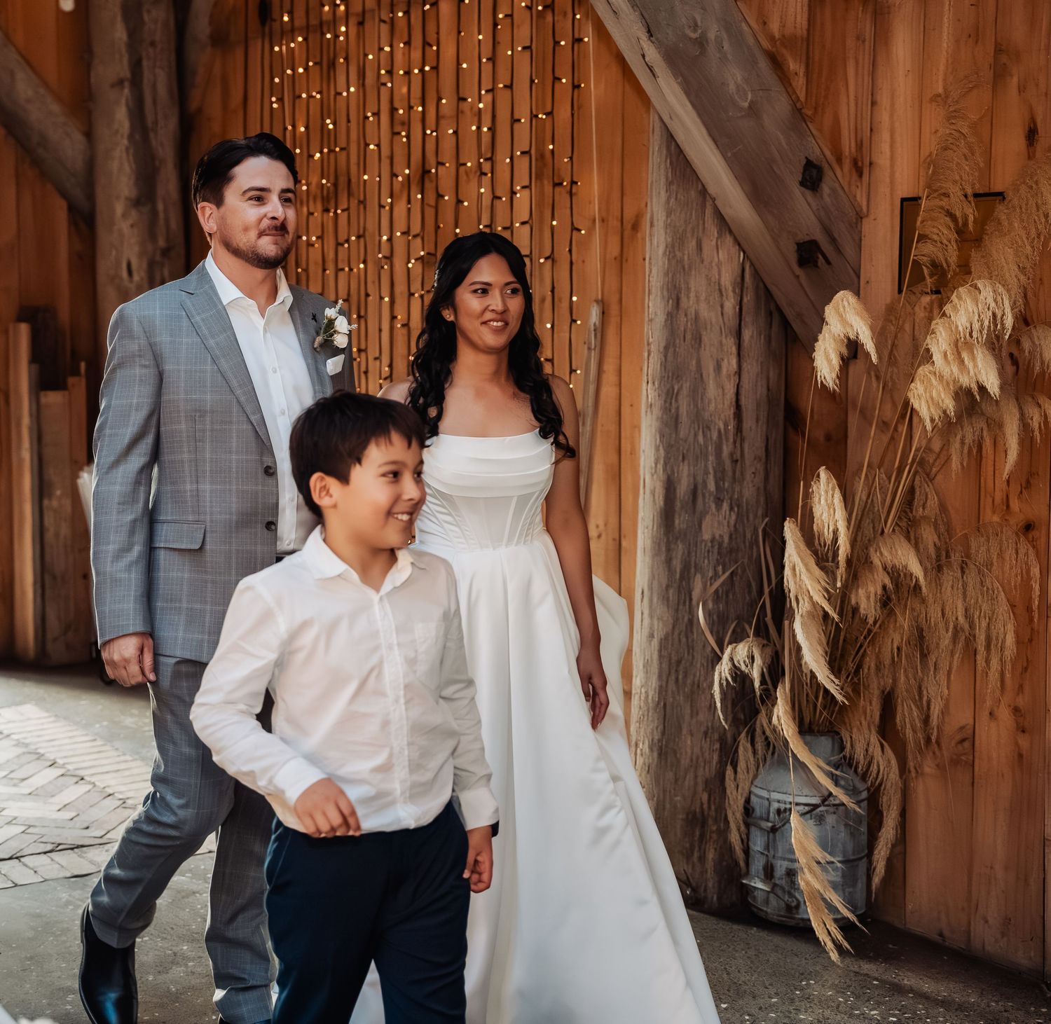 Bride in white gown and groom in gray suit walk with young boy inside a barn with fairy lights.