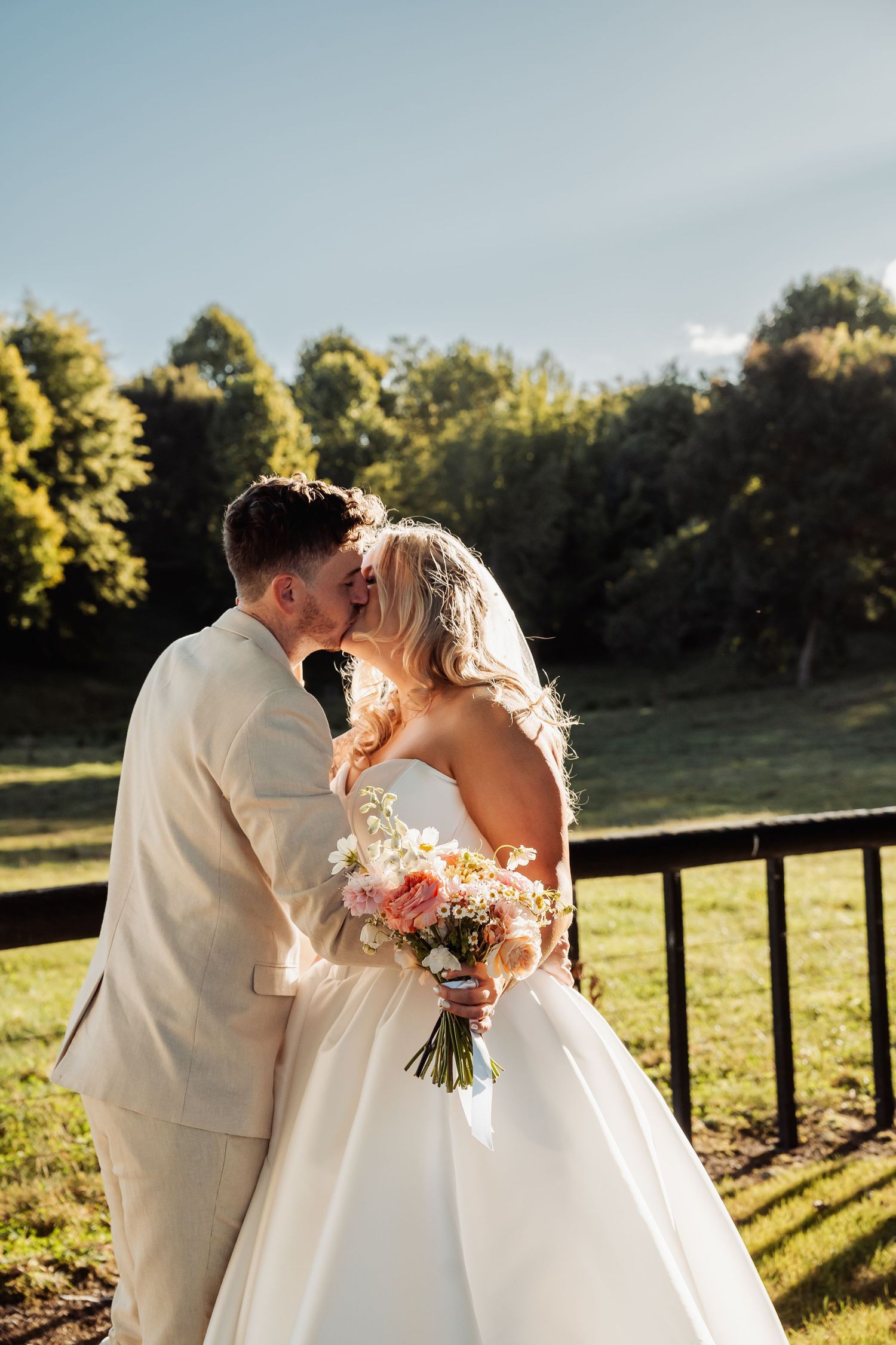 Bride and groom share a kiss outdoors in golden sunlight, bride holding a pastel bouquet.