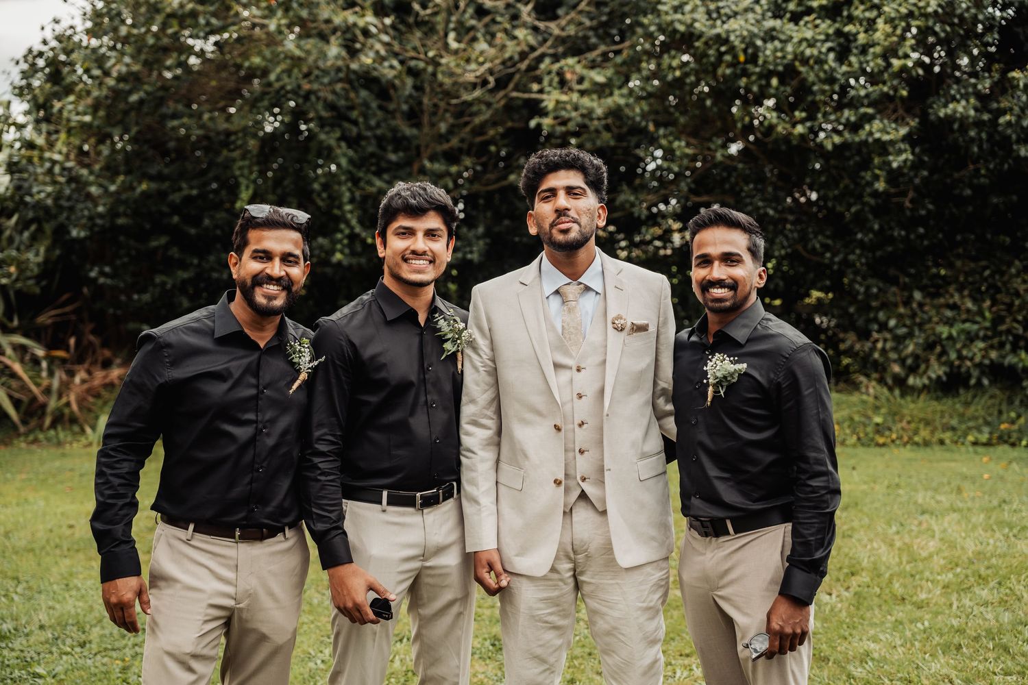 Groom in light suit poses with three groomsmen in black shirts and khaki pants outdoors.