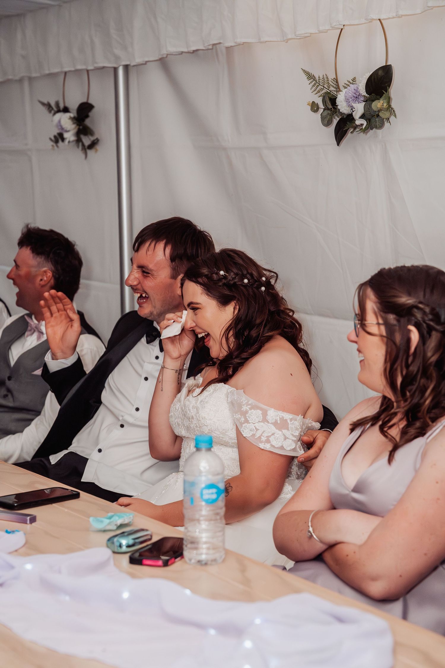 Bride laughing emotionally with groom and guests during wedding reception speeches at a decorated table.
