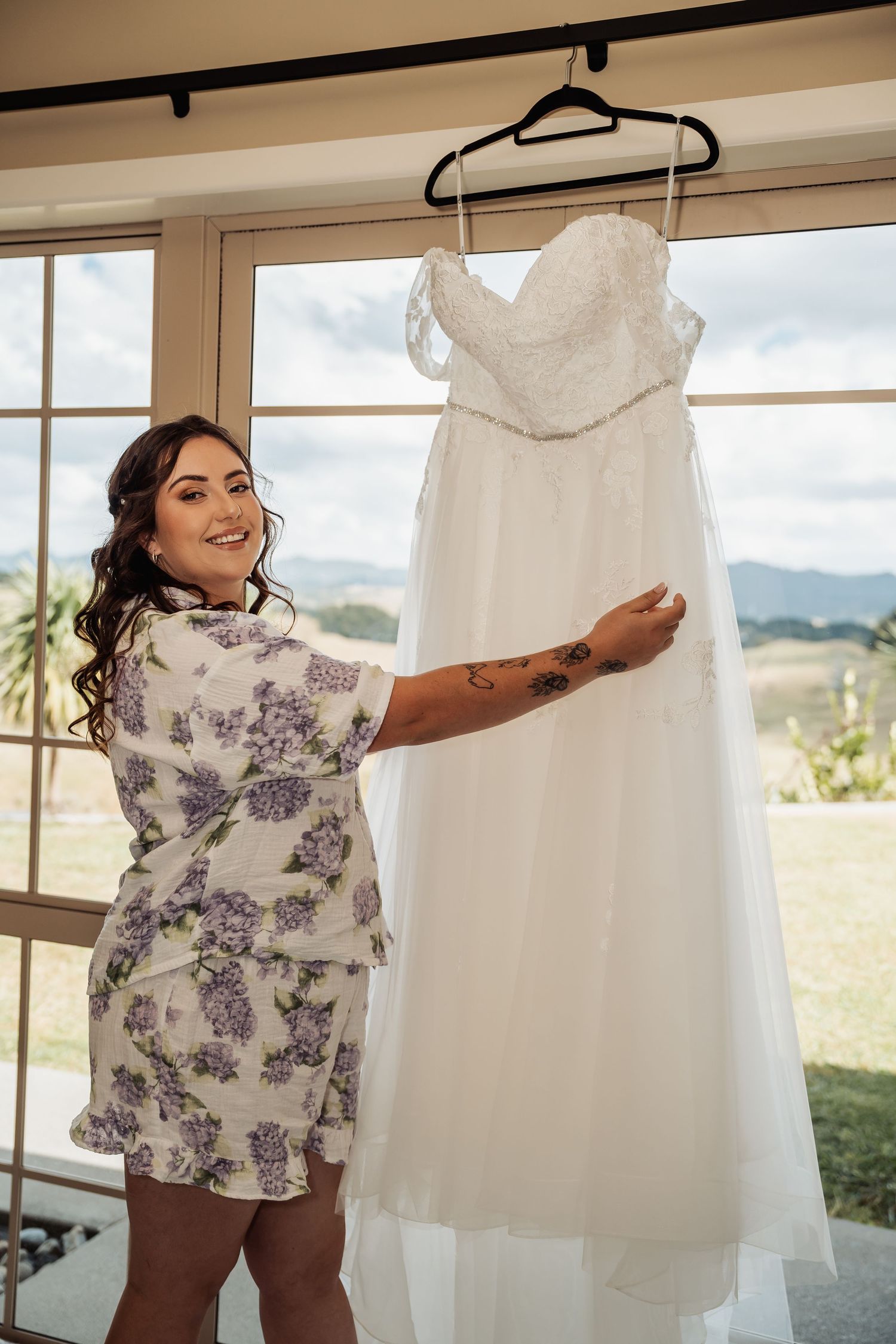 Bride smiling in floral robe holding her white lace wedding dress on a hanger by a window.