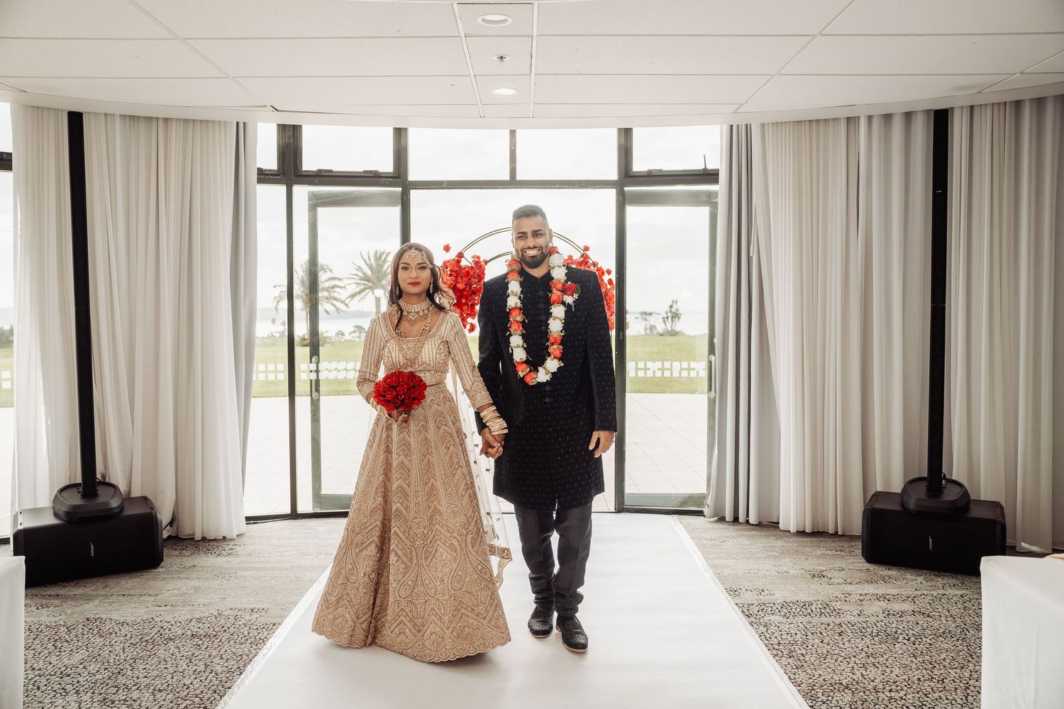 A couple walks together through an elegant white room with floor-to-ceiling windows and curtains.