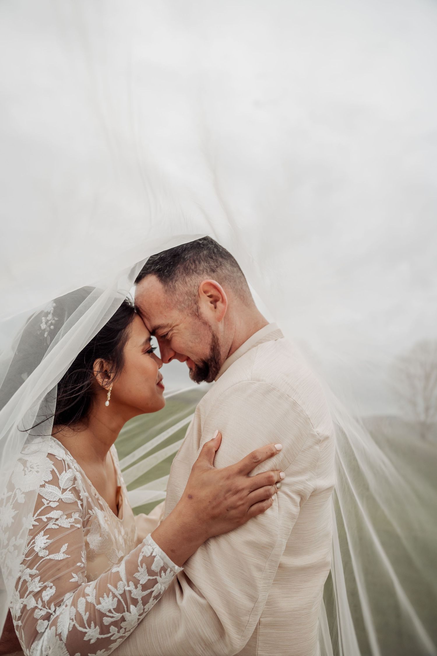 Intimate wedding couple moment under flowing veil with soft lighting.