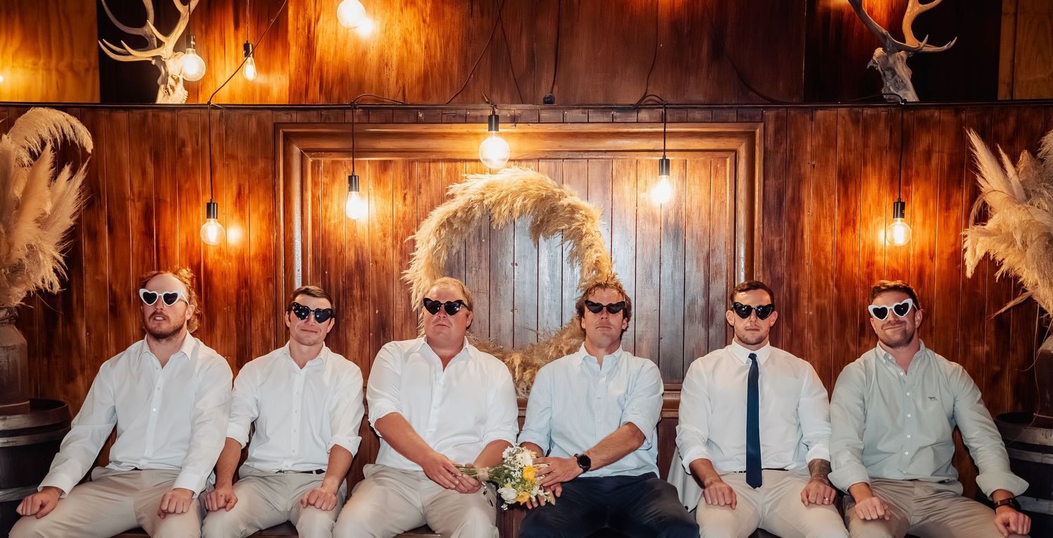 Six groomsmen in white shirts and sunglasses pose together in rustic barn venue.