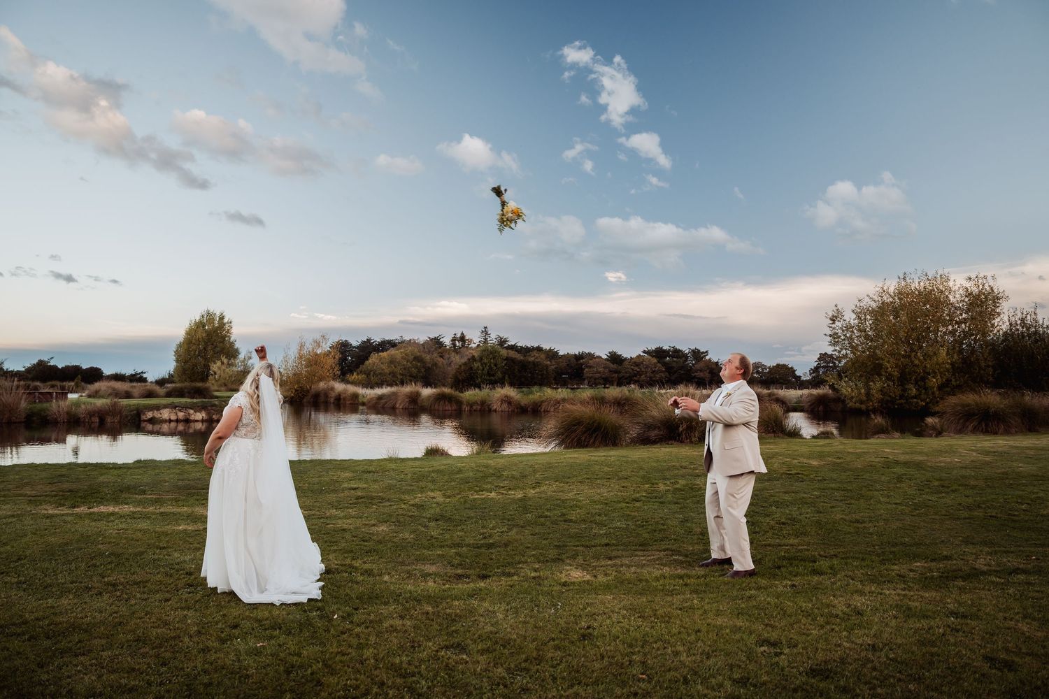 Bride and groom tossing bouquet by lakeside at sunset.