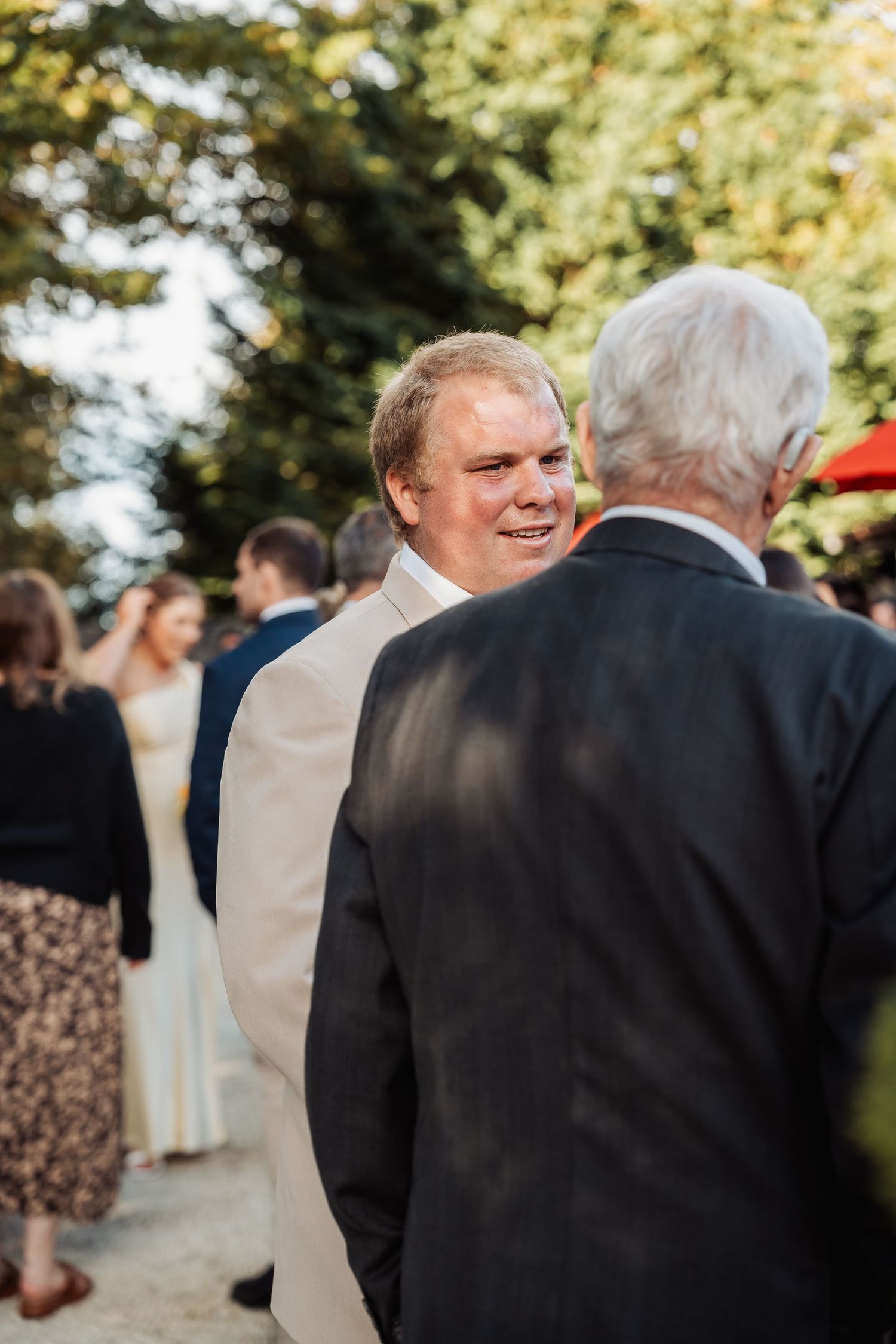 Groom in beige suit smiles while chatting with an older gentleman at an outdoor wedding reception.