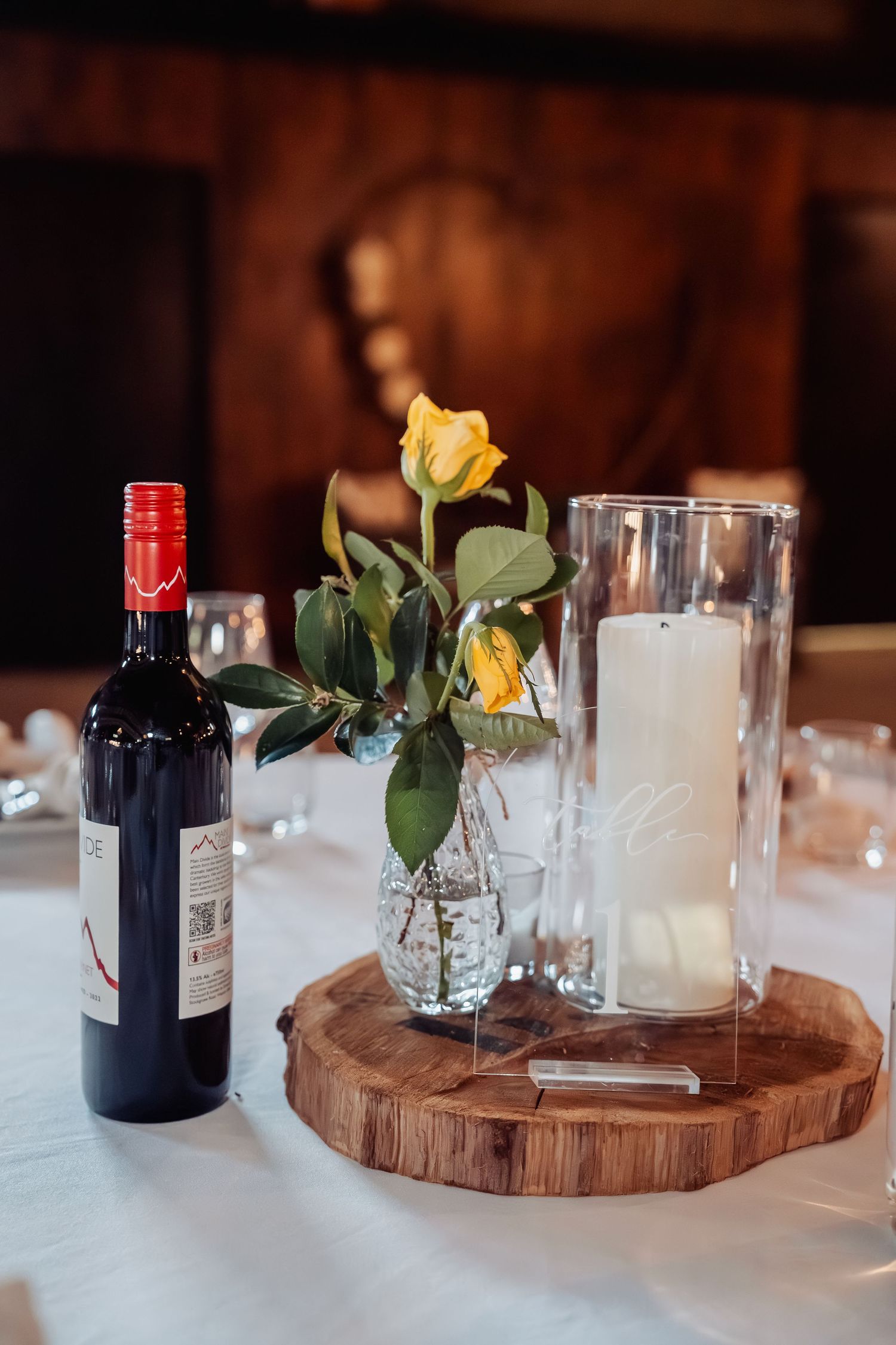Wedding table centerpiece with yellow roses, candle, and red wine bottle.