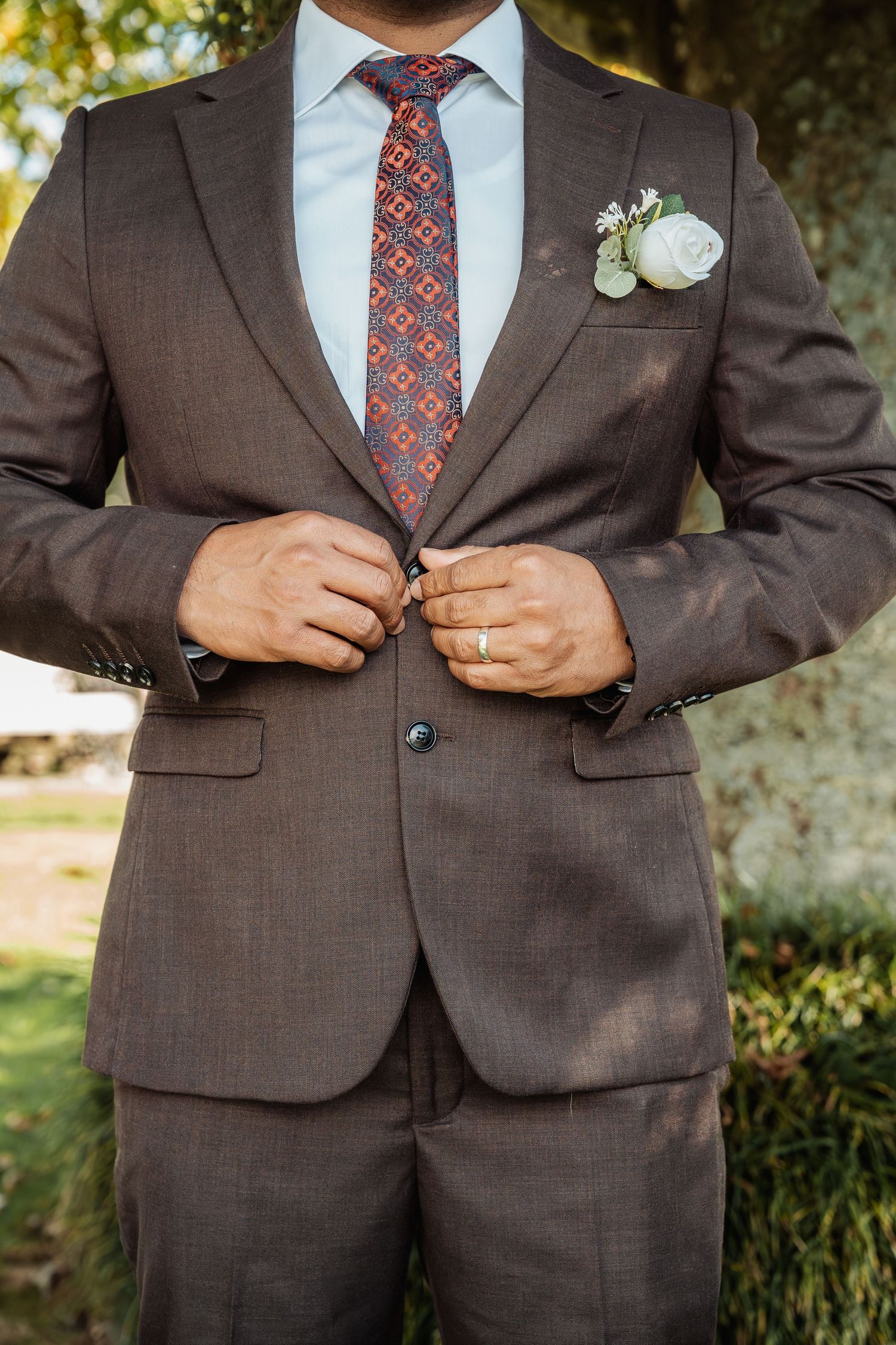 Groom in brown suit with floral boutonniere and patterned tie buttoning jacket outdoors.