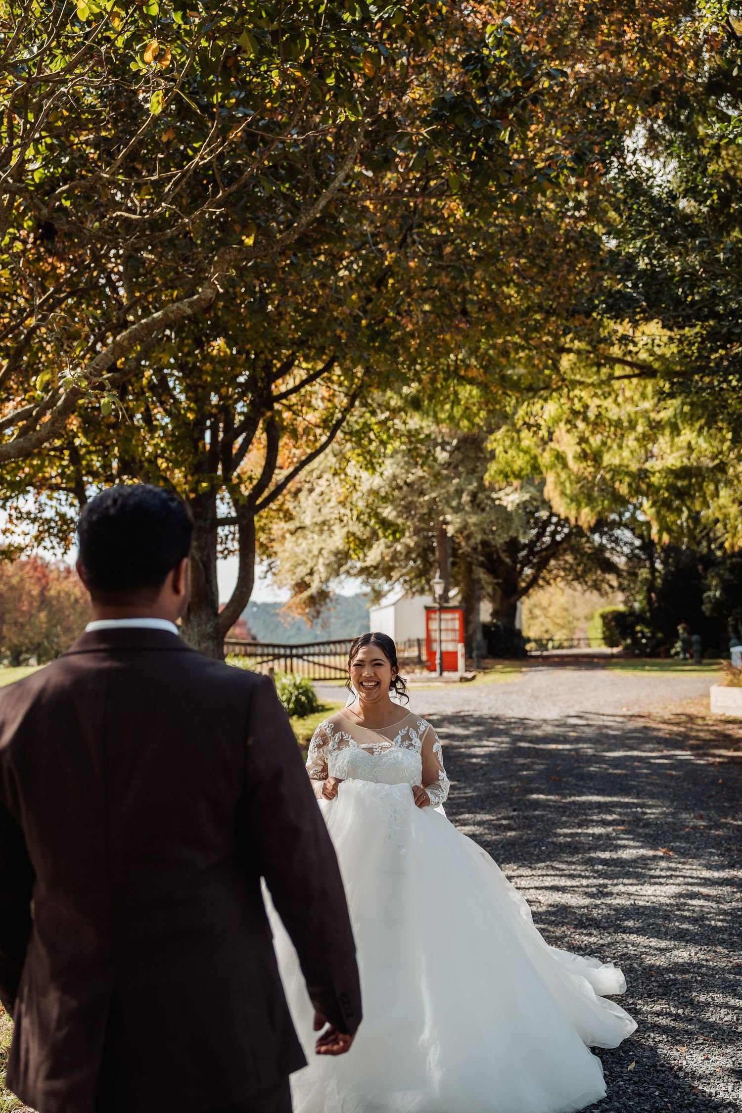 Bride in lace ball gown laughing as groom walks toward her on tree-lined autumn driveway.