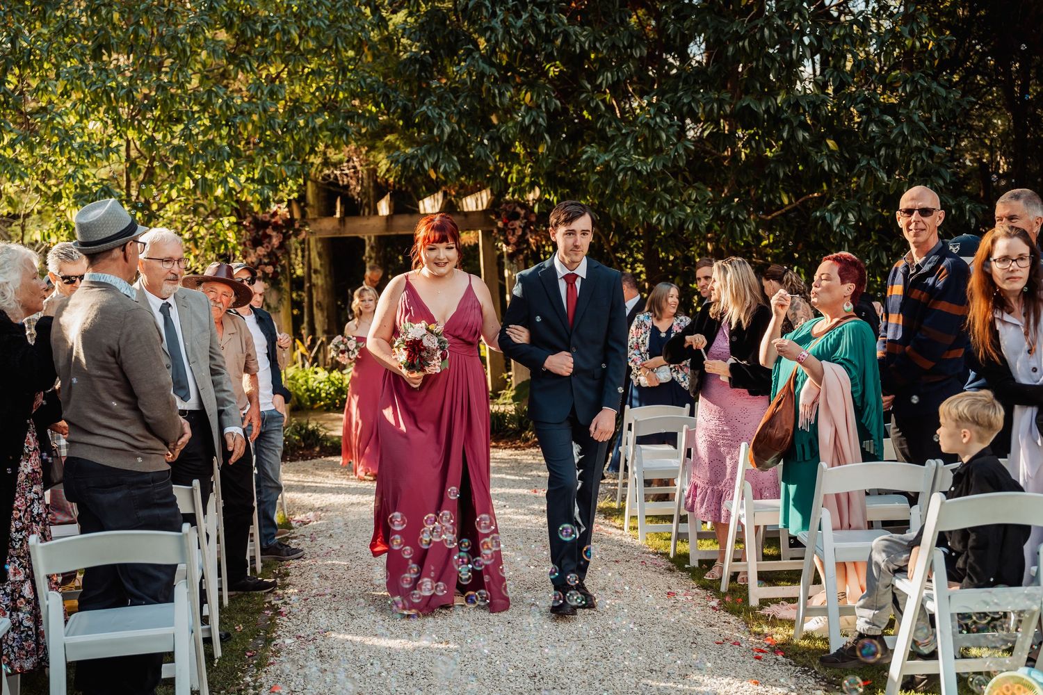 Bridesmaid in burgundy gown walks down outdoor aisle with groomsman past seated wedding guests.