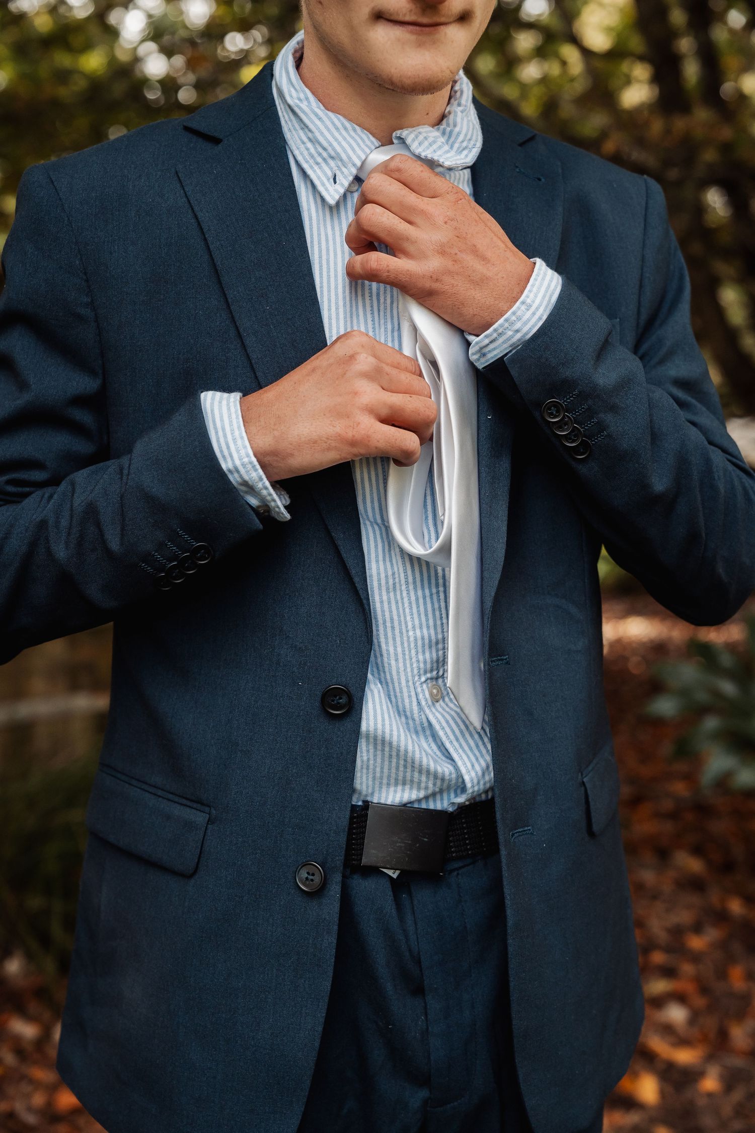 Groom in navy suit adjusting white tie outdoors among autumn foliage.