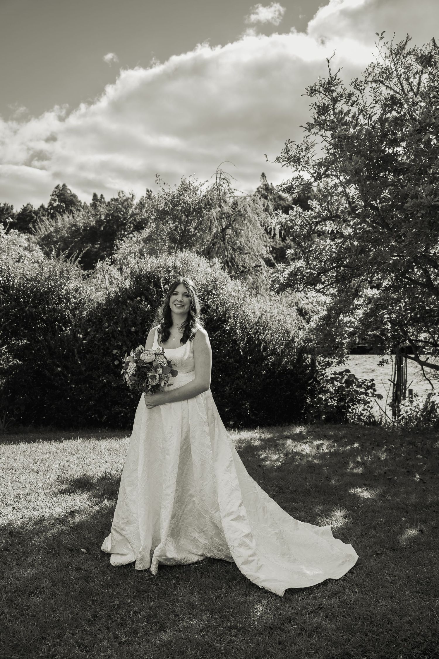 Black and white portrait of bride in flowing white gown holding bouquet in garden.