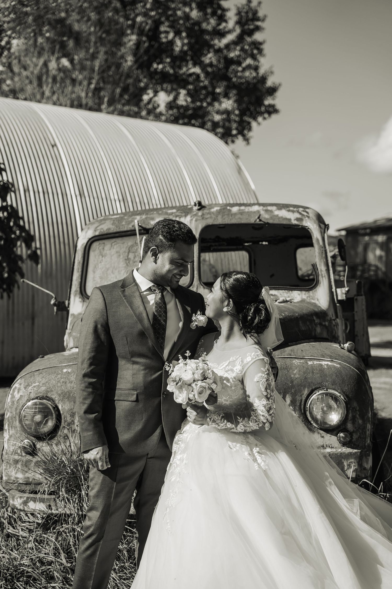 Black and white wedding portrait of couple embracing beside a vintage rustic truck.