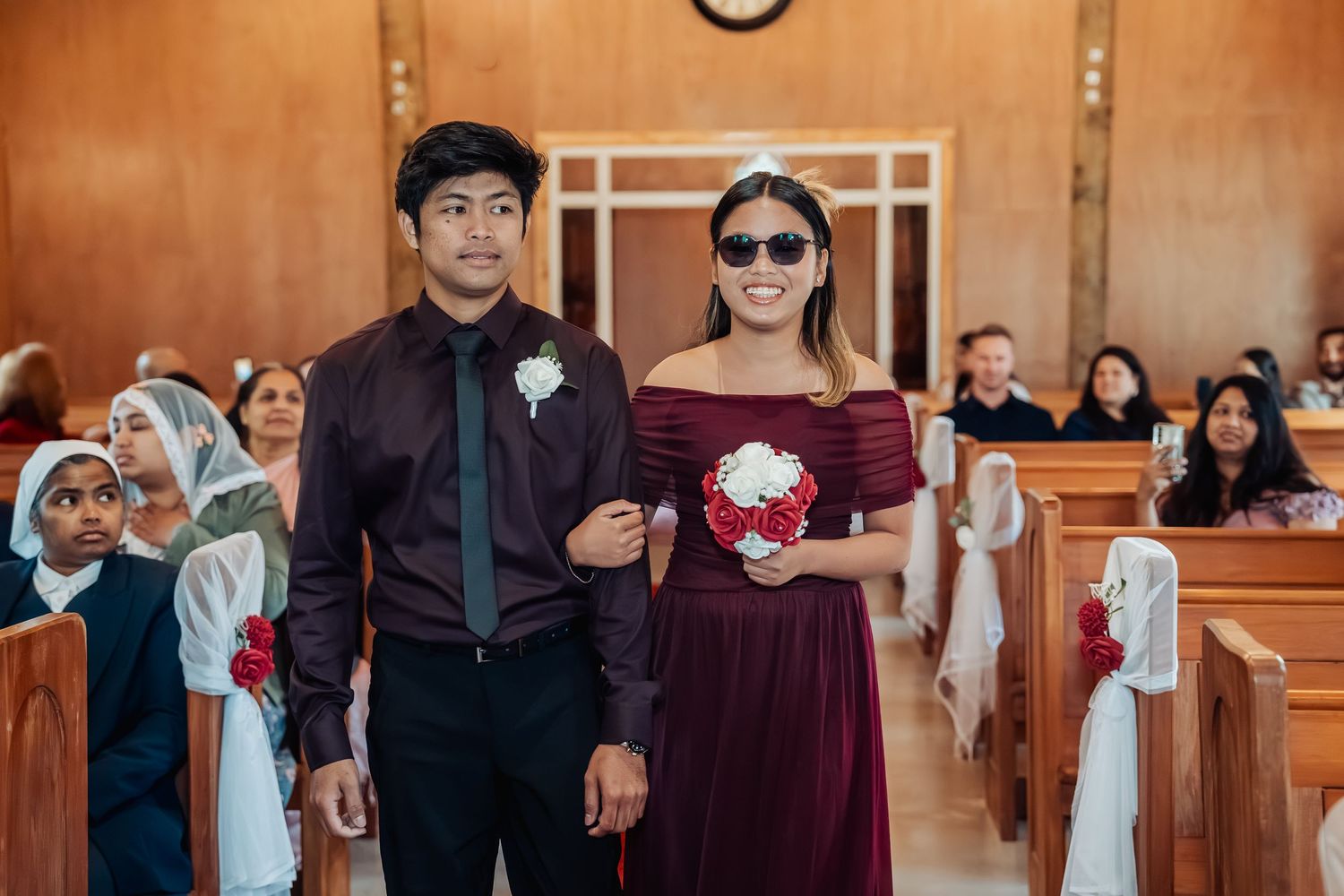 A groomsman in burgundy and a bridesmaid in a matching gown walk down a church aisle holding a red bouquet.