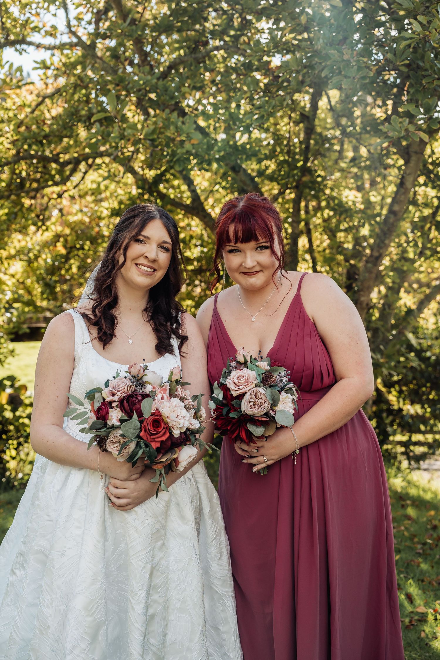 Bride in white lace gown poses with bridesmaid in burgundy dress holding bouquets in garden.