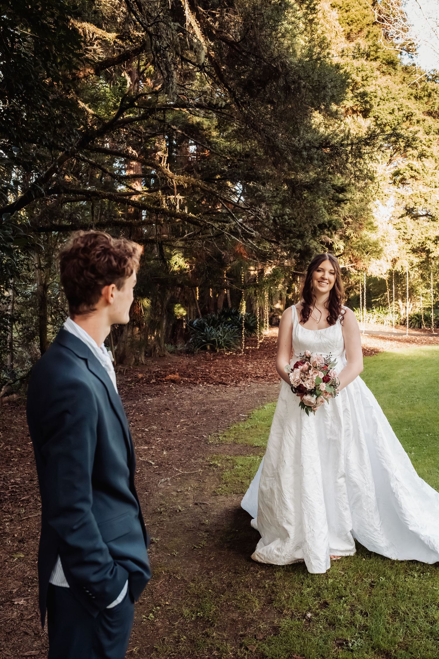 Bride in white gown holding bouquet faces groom in navy suit in forest setting.
