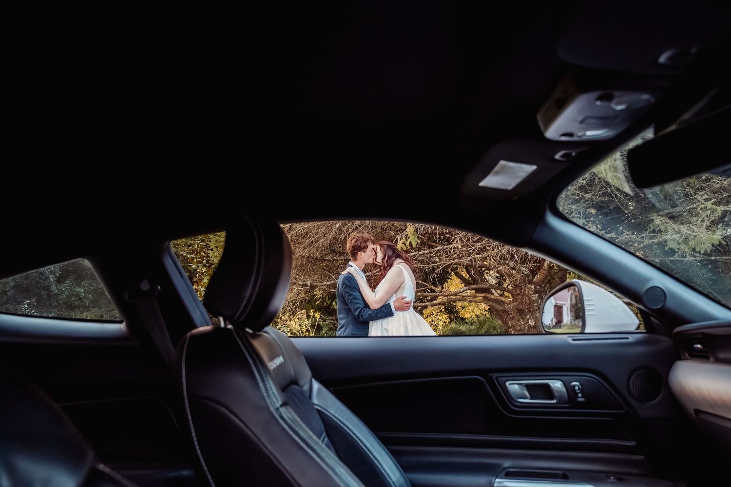 Bride and groom share a kiss viewed through the interior of a luxury car window.