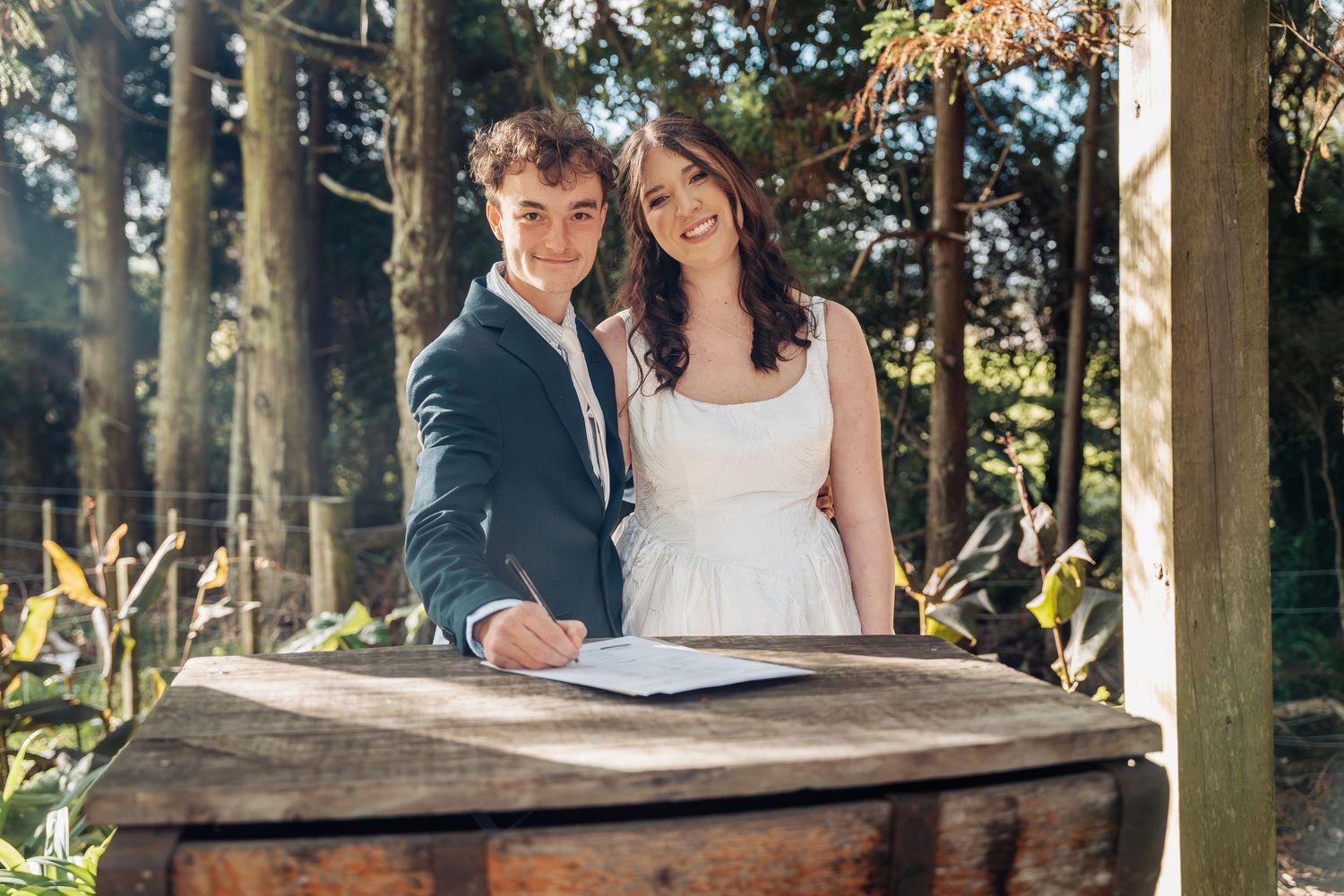 Smiling newlyweds signing wedding register at rustic wooden table in sunlit garden setting.