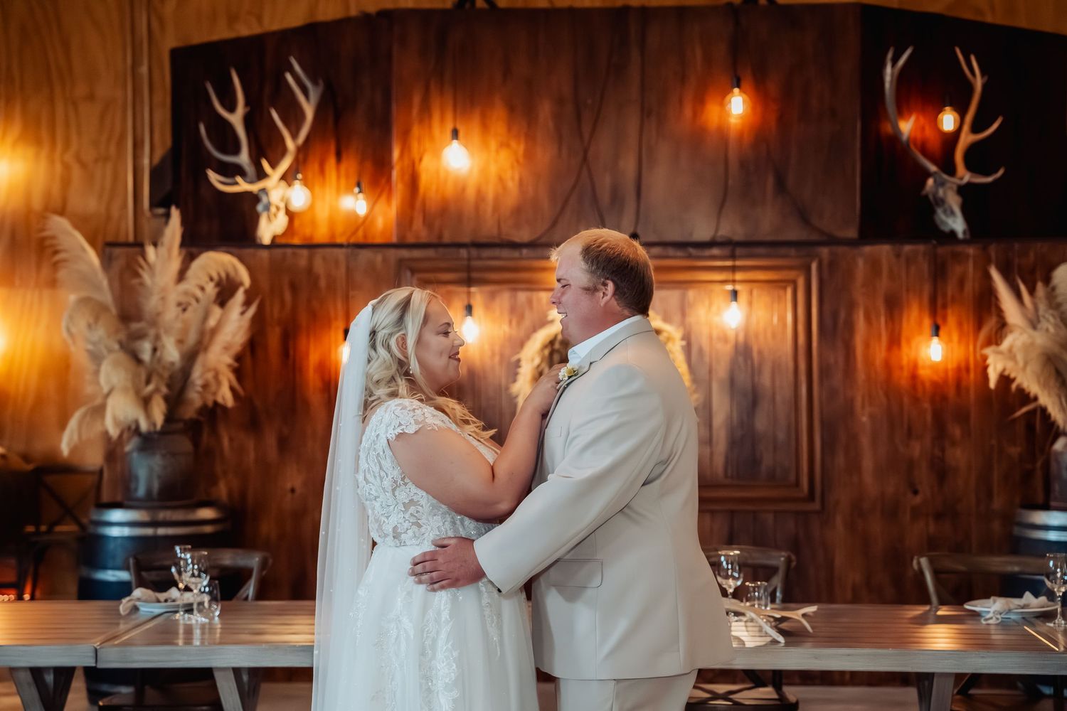 Bride and groom share a romantic first dance in a rustic barn venue with warm Edison bulb lighting and antler decor.