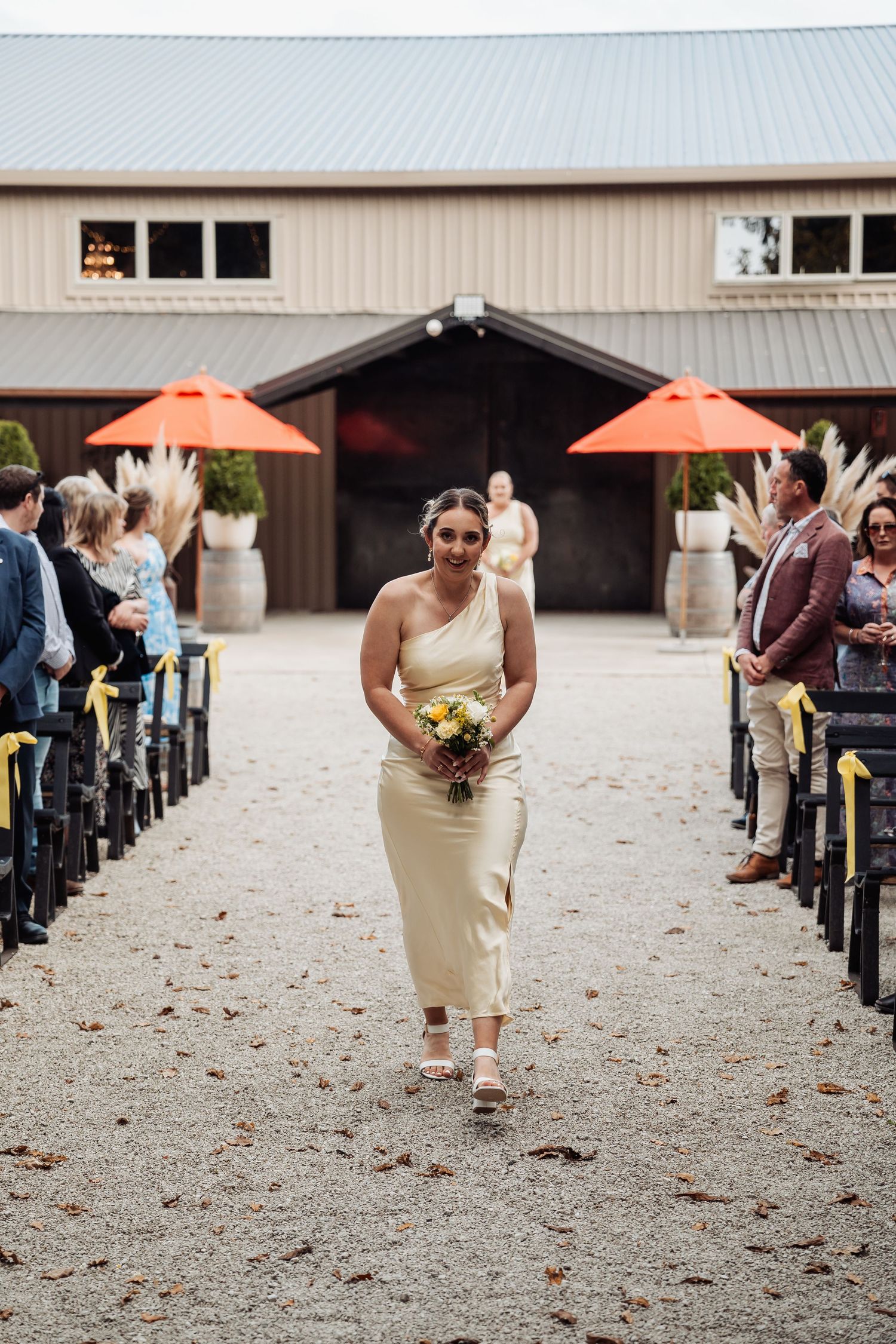 Bridesmaid walks down outdoor wedding aisle in cream dress carrying yellow and white bouquet.