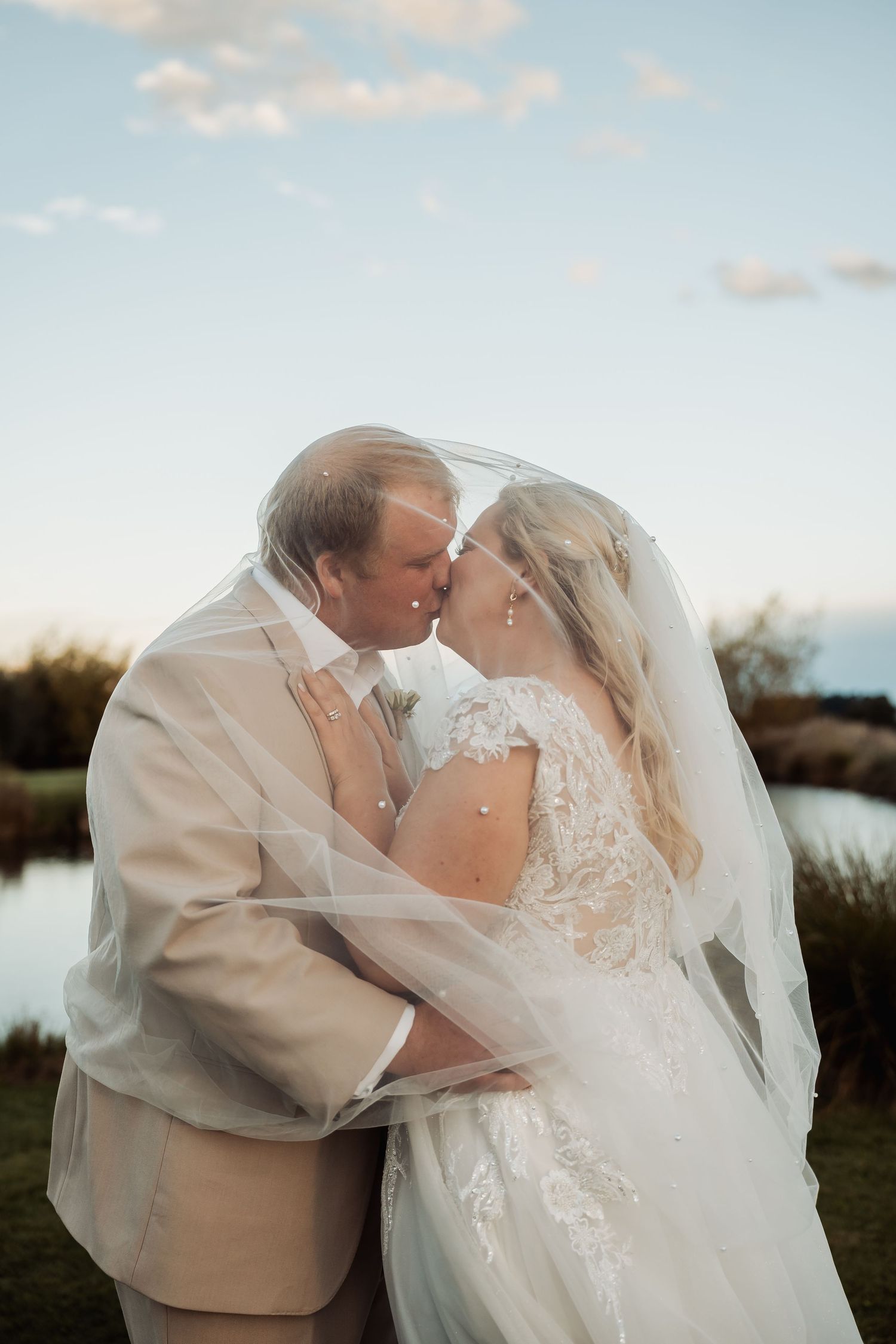 Bride and groom share a romantic kiss under flowing veil near a pond at sunset.