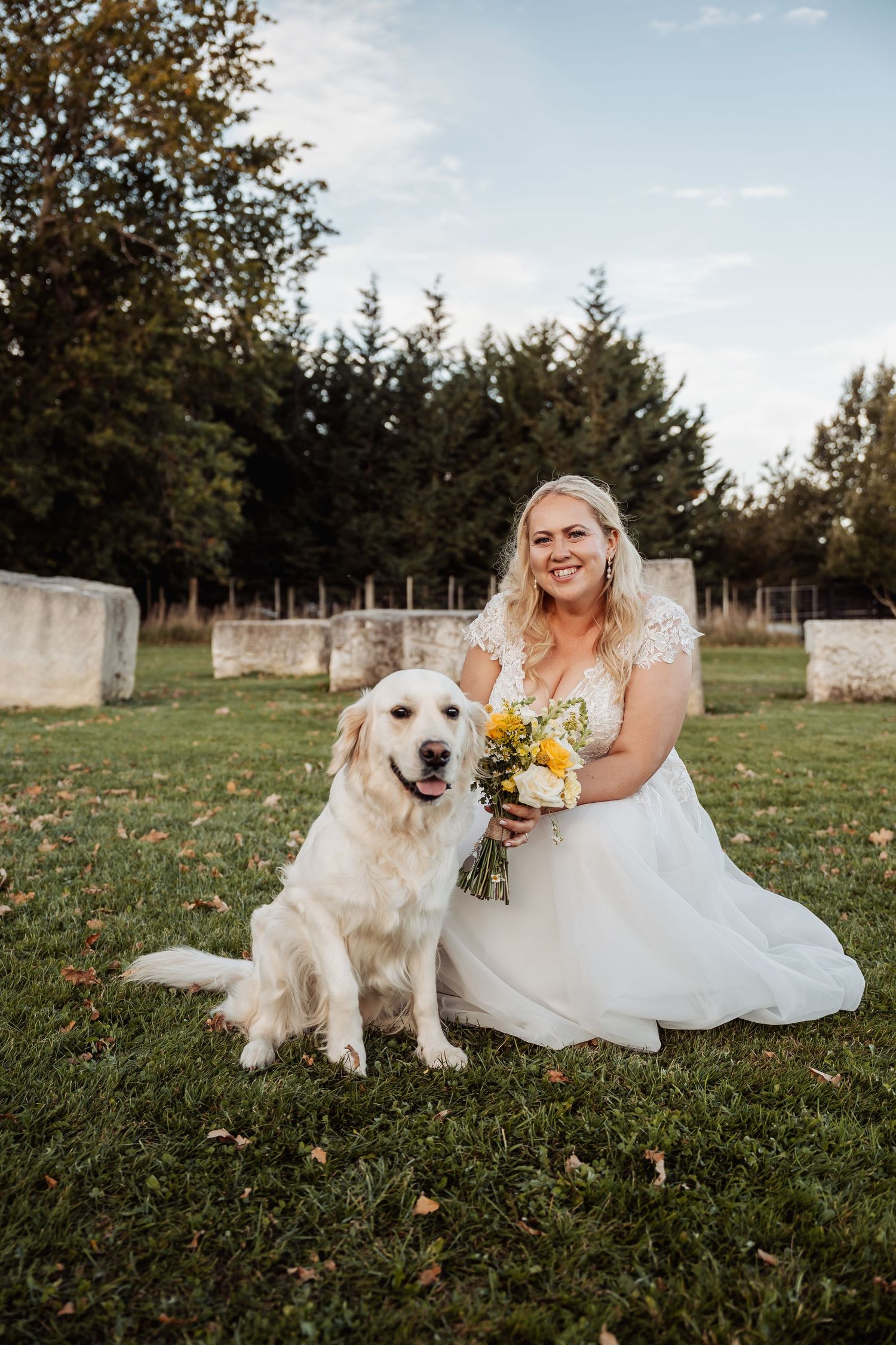 Smiling bride in lace gown poses with golden retriever dog while holding yellow bouquet.