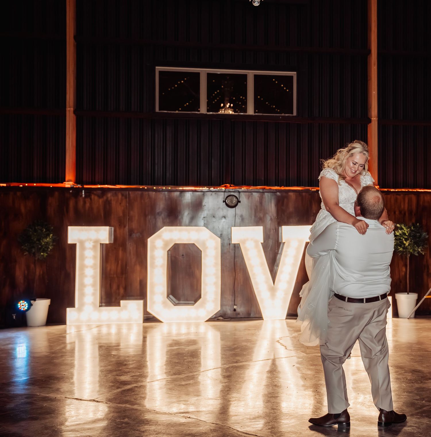 Bride and groom share first dance in front of illuminated LOVE marquee letters in barn.
