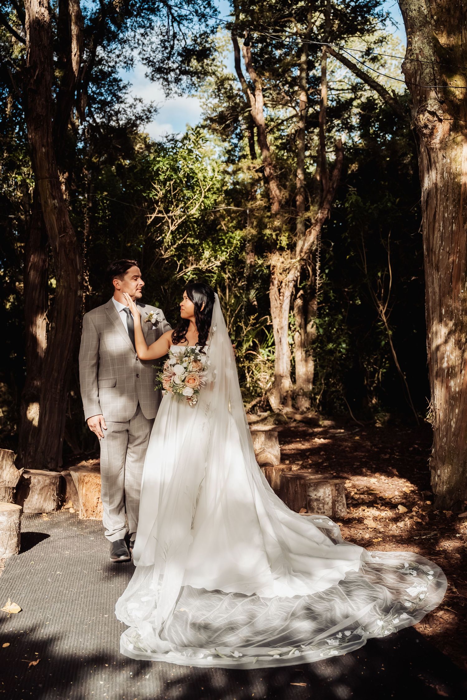 Newlyweds walk hand-in-hand along sunlit forest path, bride in flowing gown with long veil.