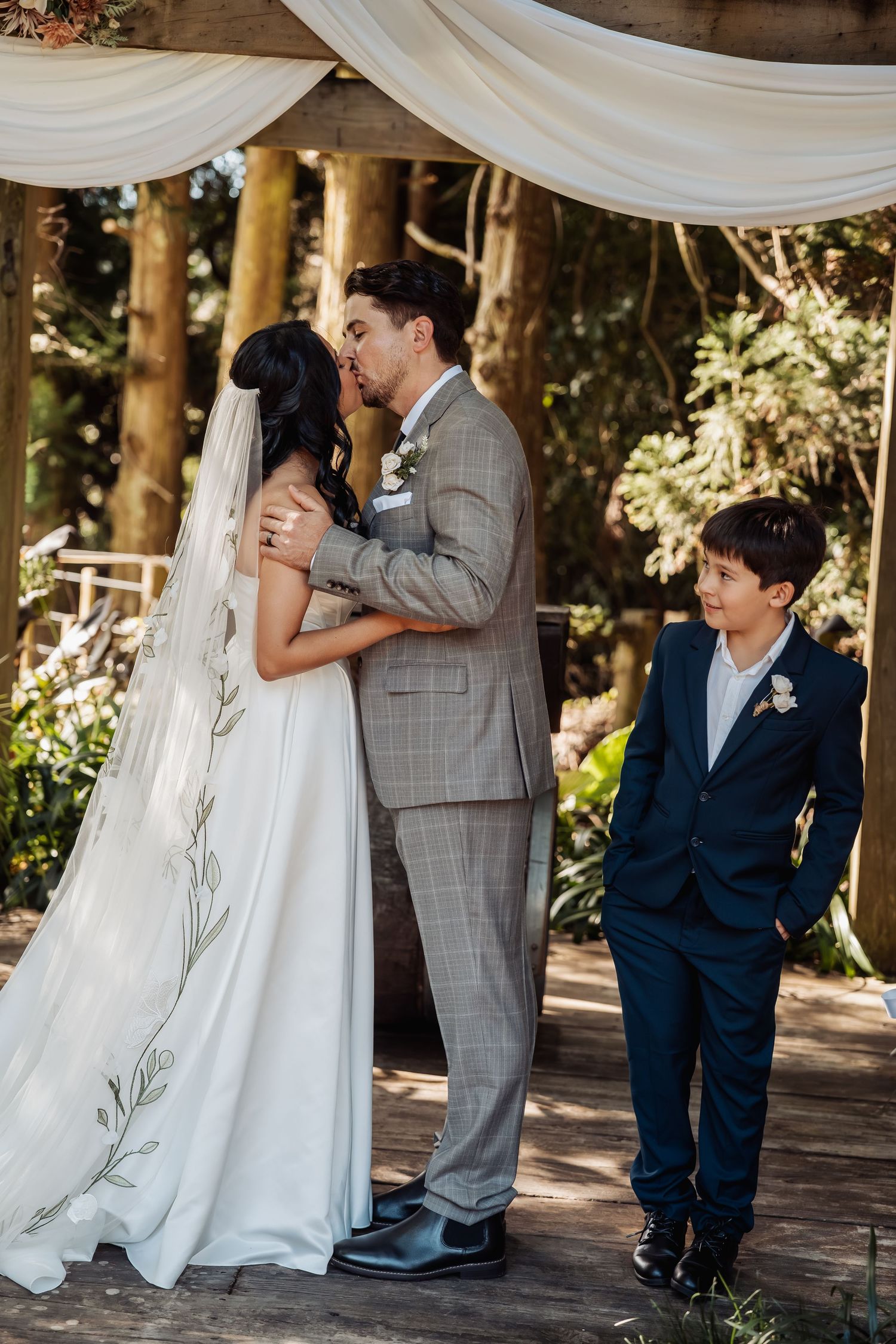 Bride and groom share first kiss at outdoor woodland ceremony while young boy watches.