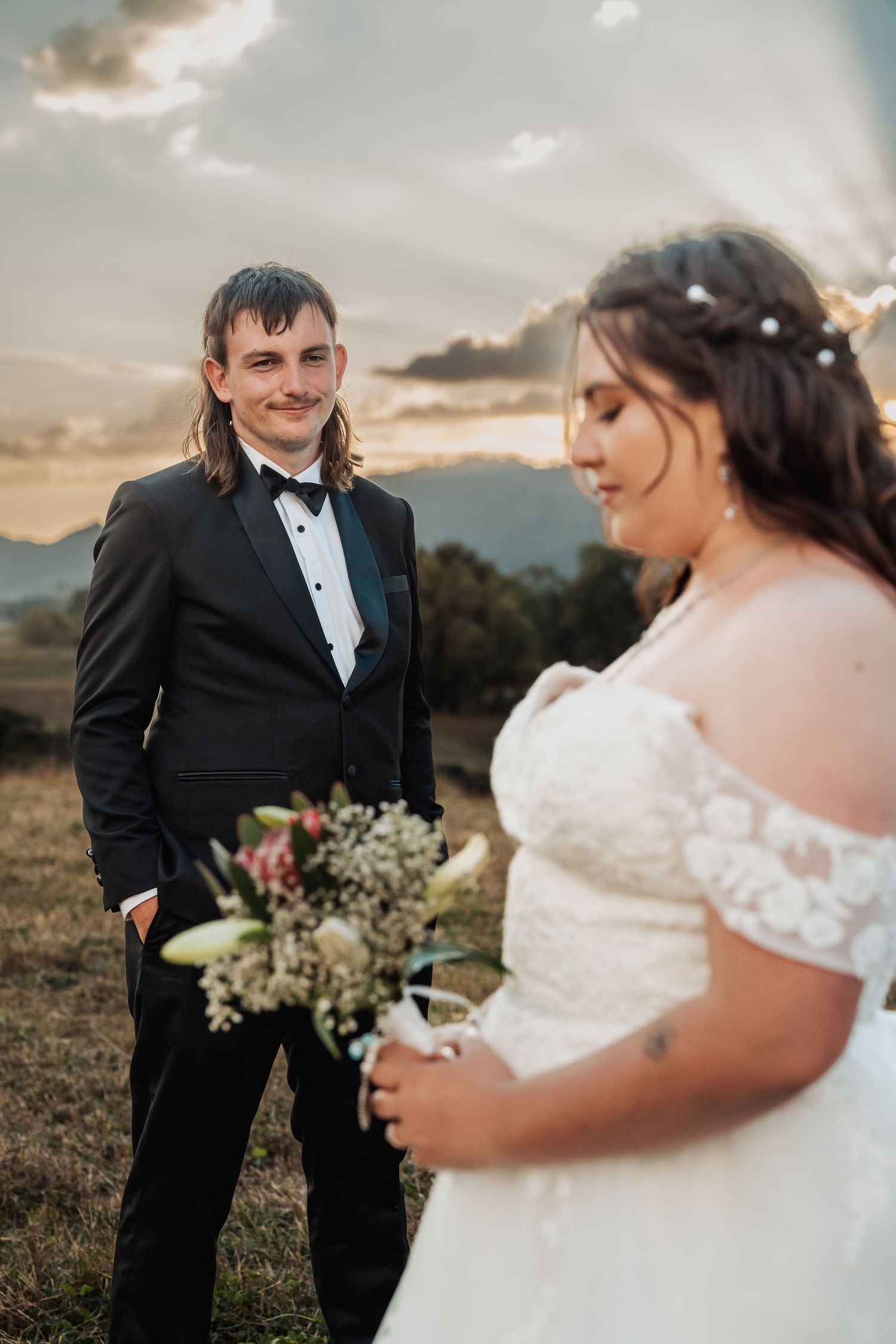 Groom in black tuxedo smiles at bride holding a bouquet during a stunning countryside sunset.