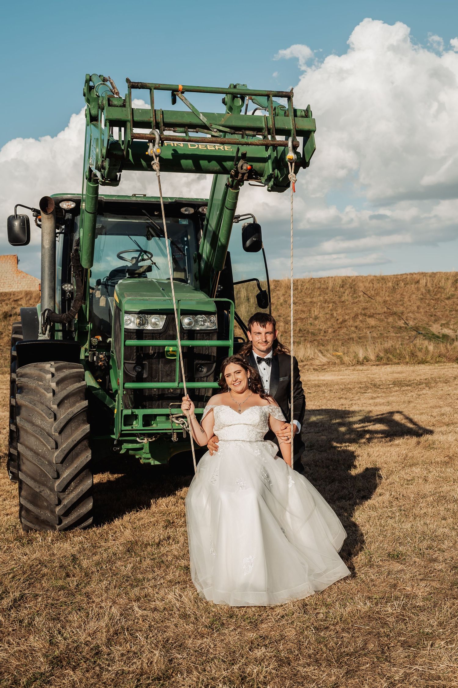 Bride and groom posing on a rope swing in front of a green John Deere tractor in a golden field.