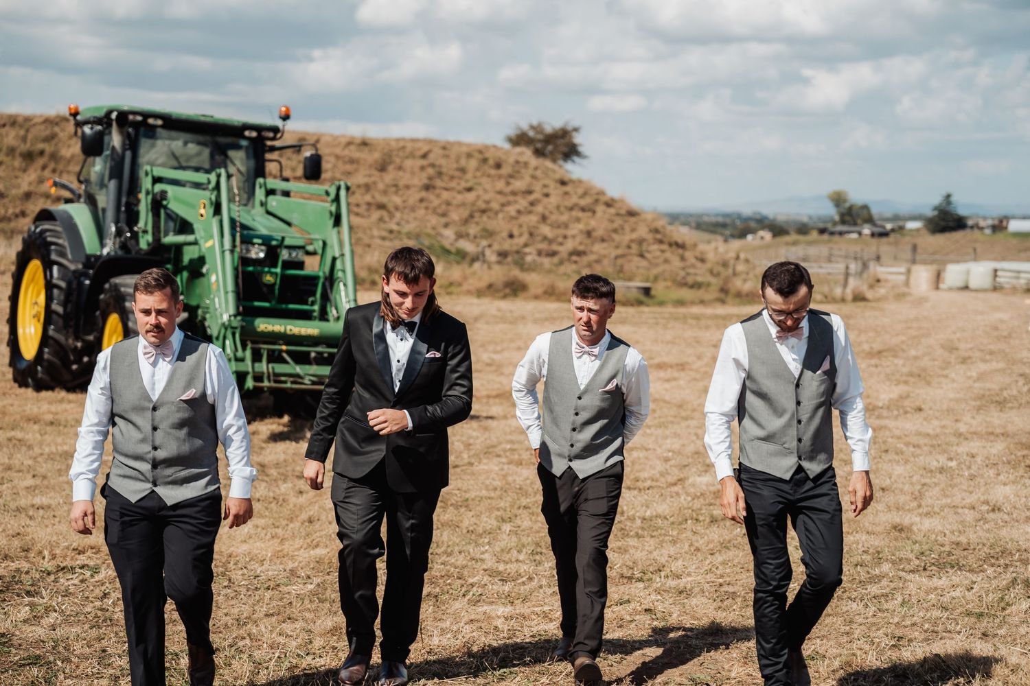 Groom and groomsmen in grey vests walk across dry field with green John Deere tractor behind them.