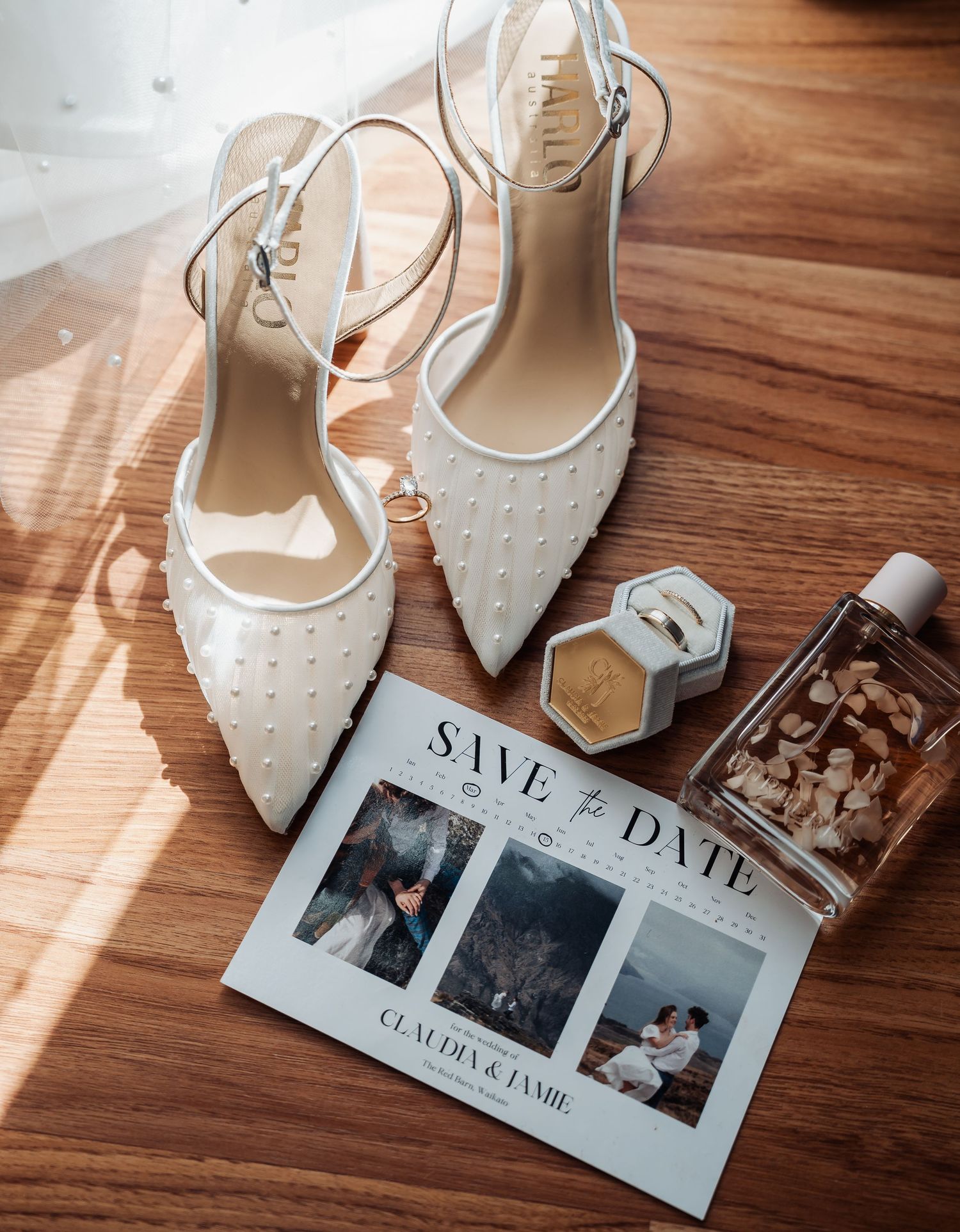 White pearl wedding heels beside a save the date card, ring box, and perfume bottle on wooden floor.