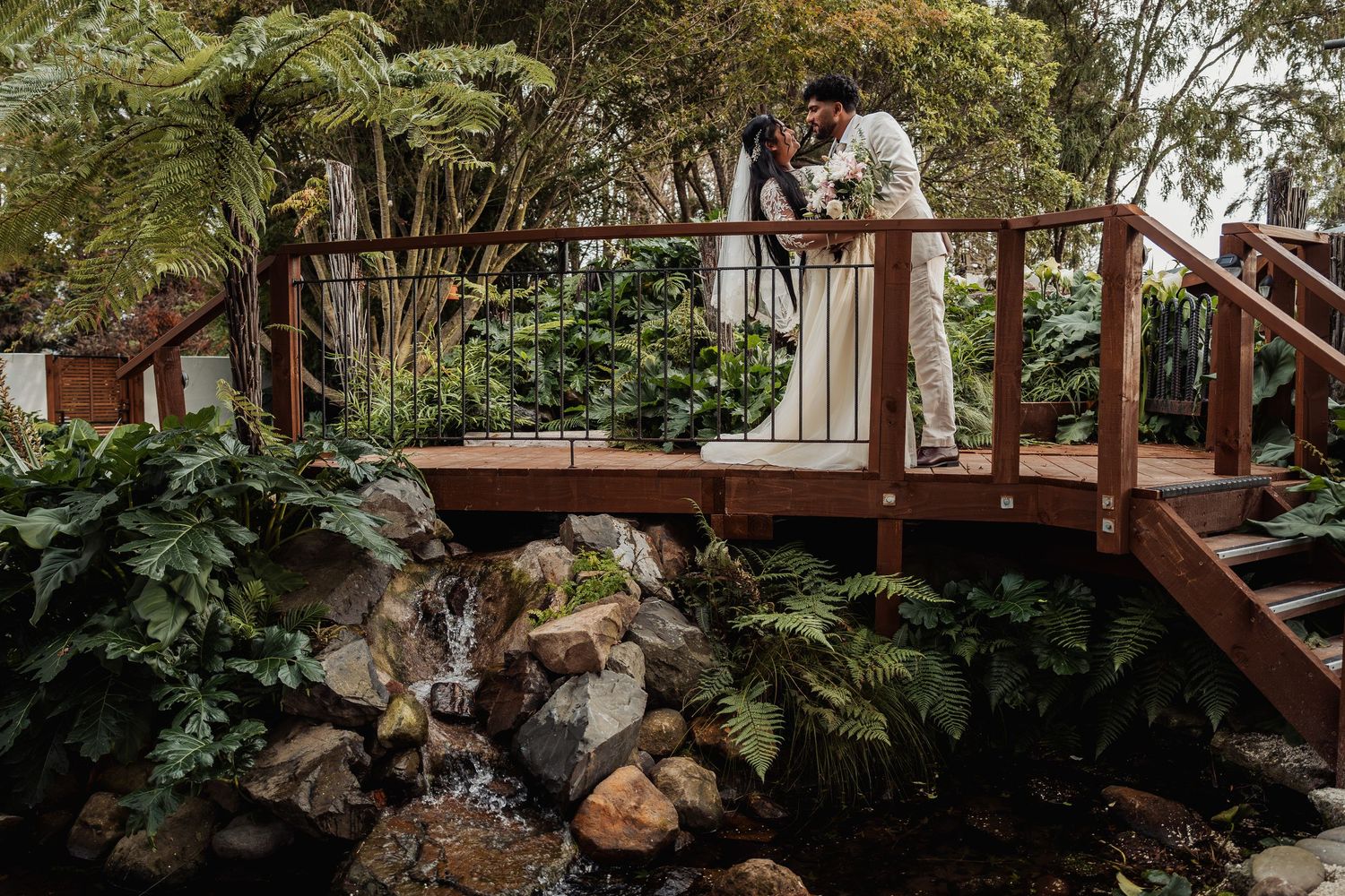 Bride and groom sharing romantic moment on wooden bridge surrounded by tropical garden waterfall.