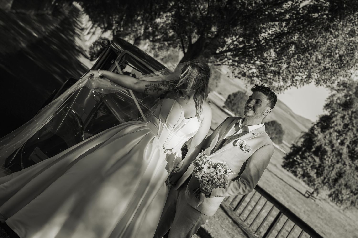 Black and white photo of bride and groom standing by vintage car under tree.