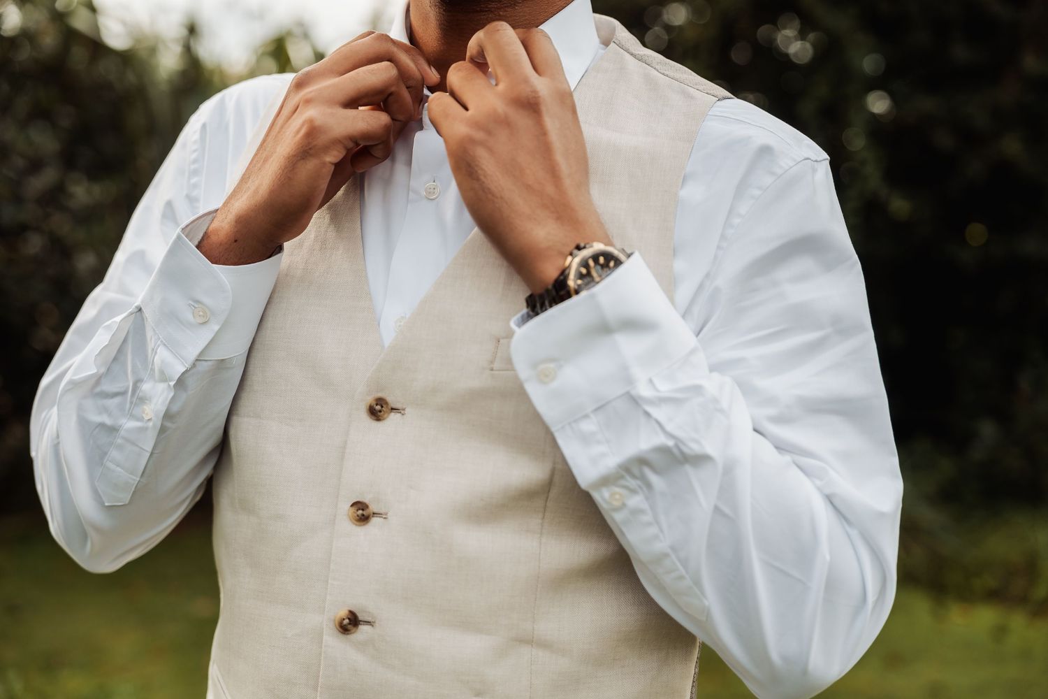 Man in white dress shirt and beige vest adjusting his tie, wearing a watch, outdoors at a wedding.